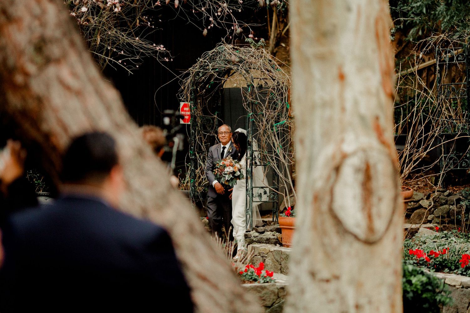 bride seen through trees walking down with father to wedding ceremony