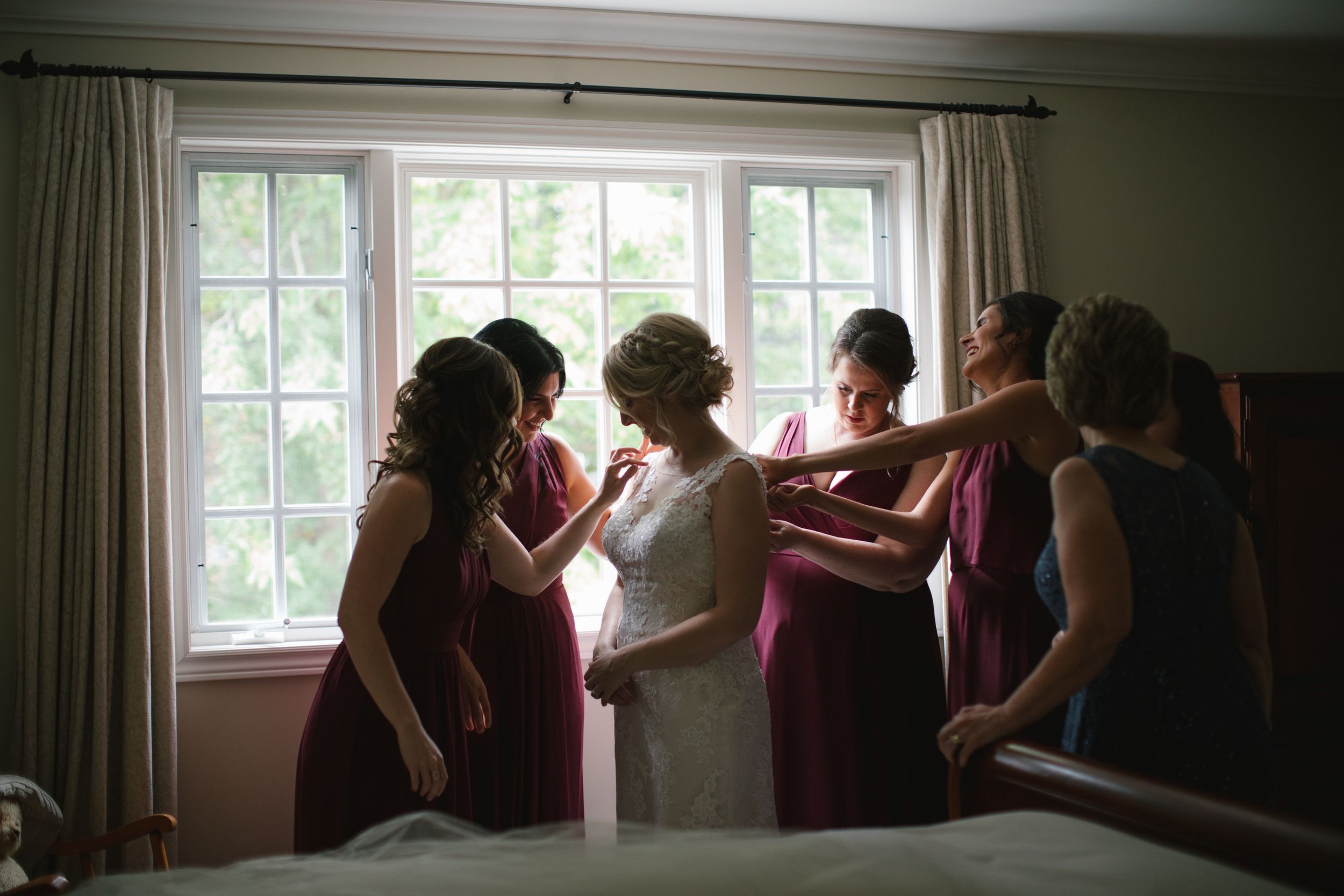 bridesmaids in burgundy dresses helping bride into her wedding dress against the window