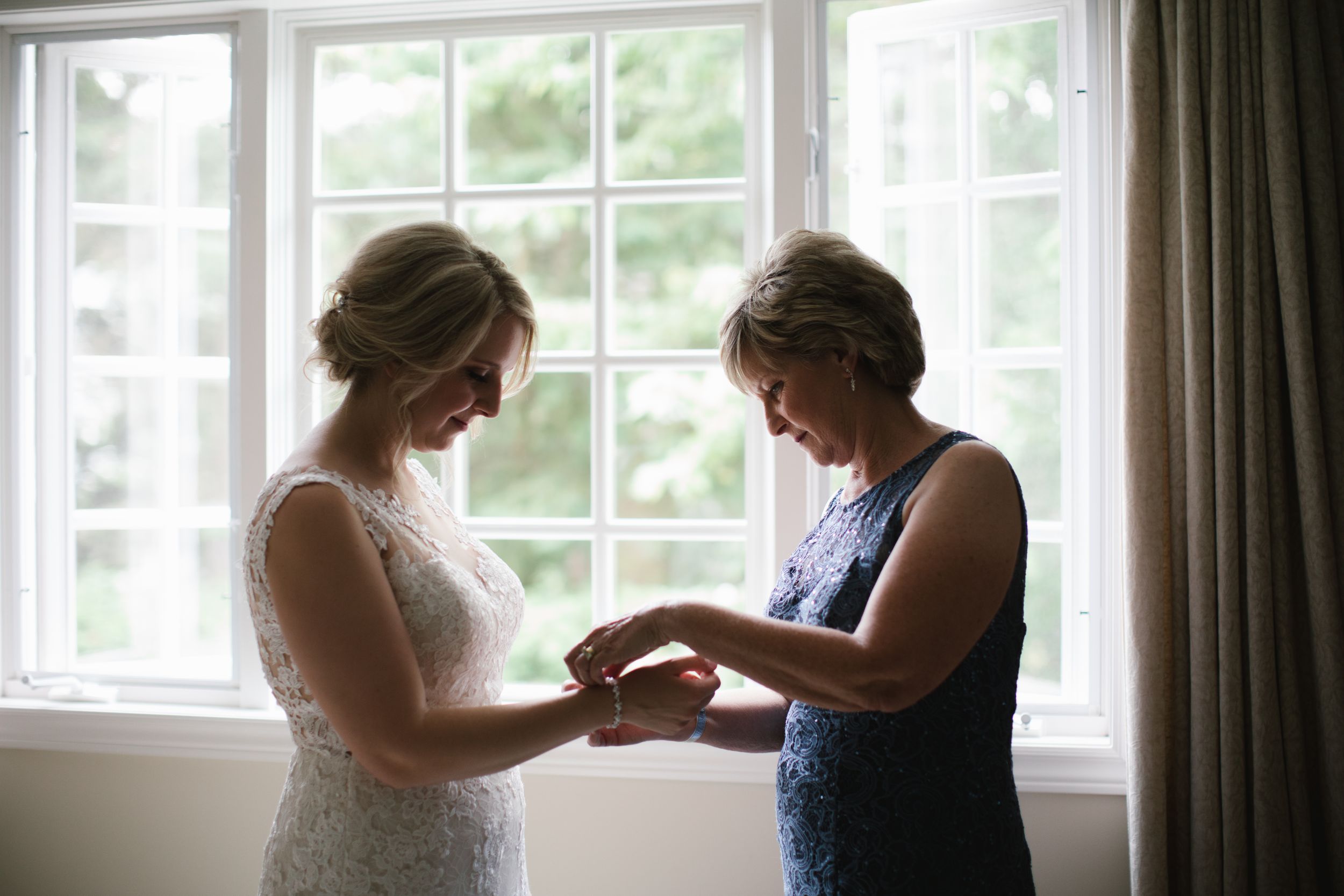 mother of bride helping bride put on her jewelry against the window
