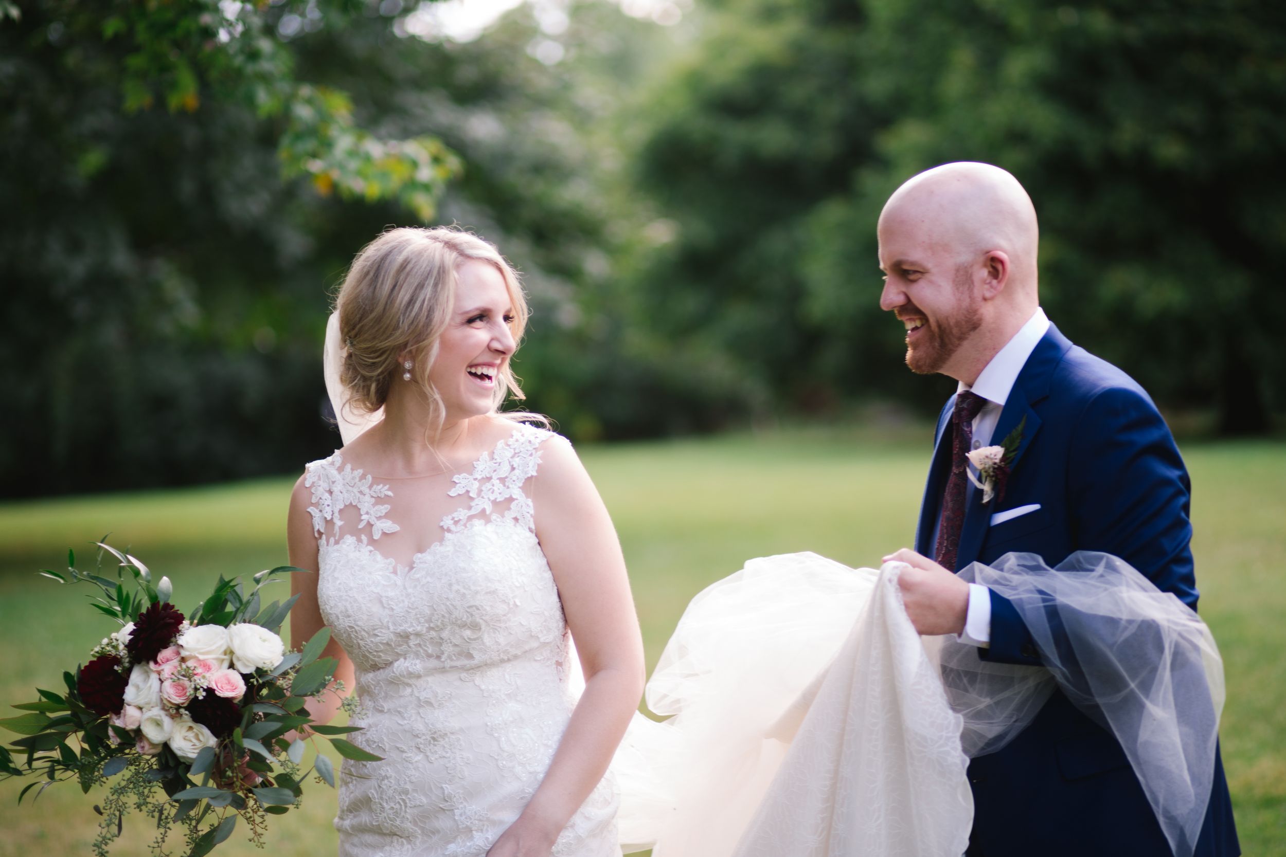 groom holding brides wedding dress and veil, looking at each other and laughing