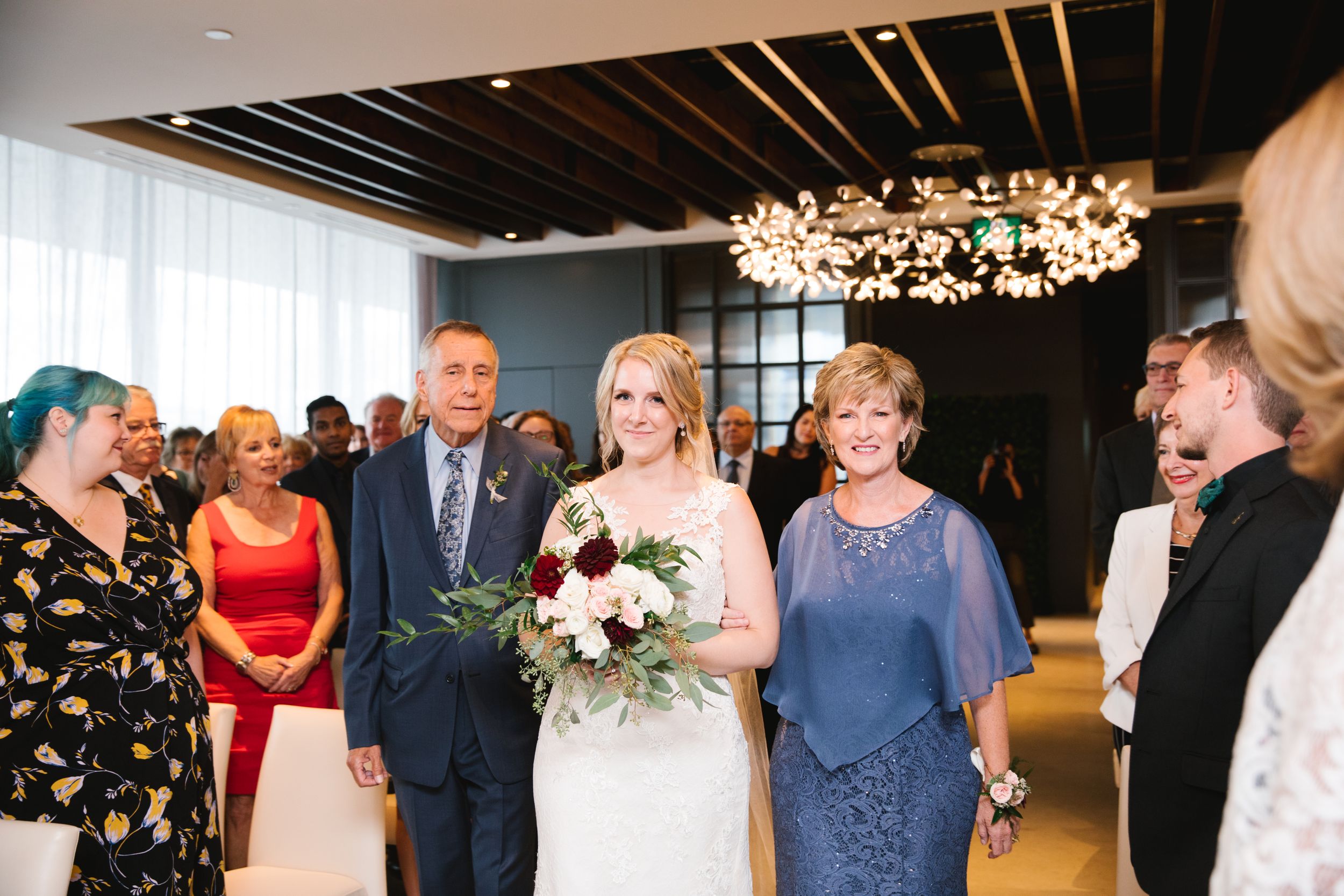 bride walking down aisle with both parents and holding a bouquet of burgundy and pink flowers