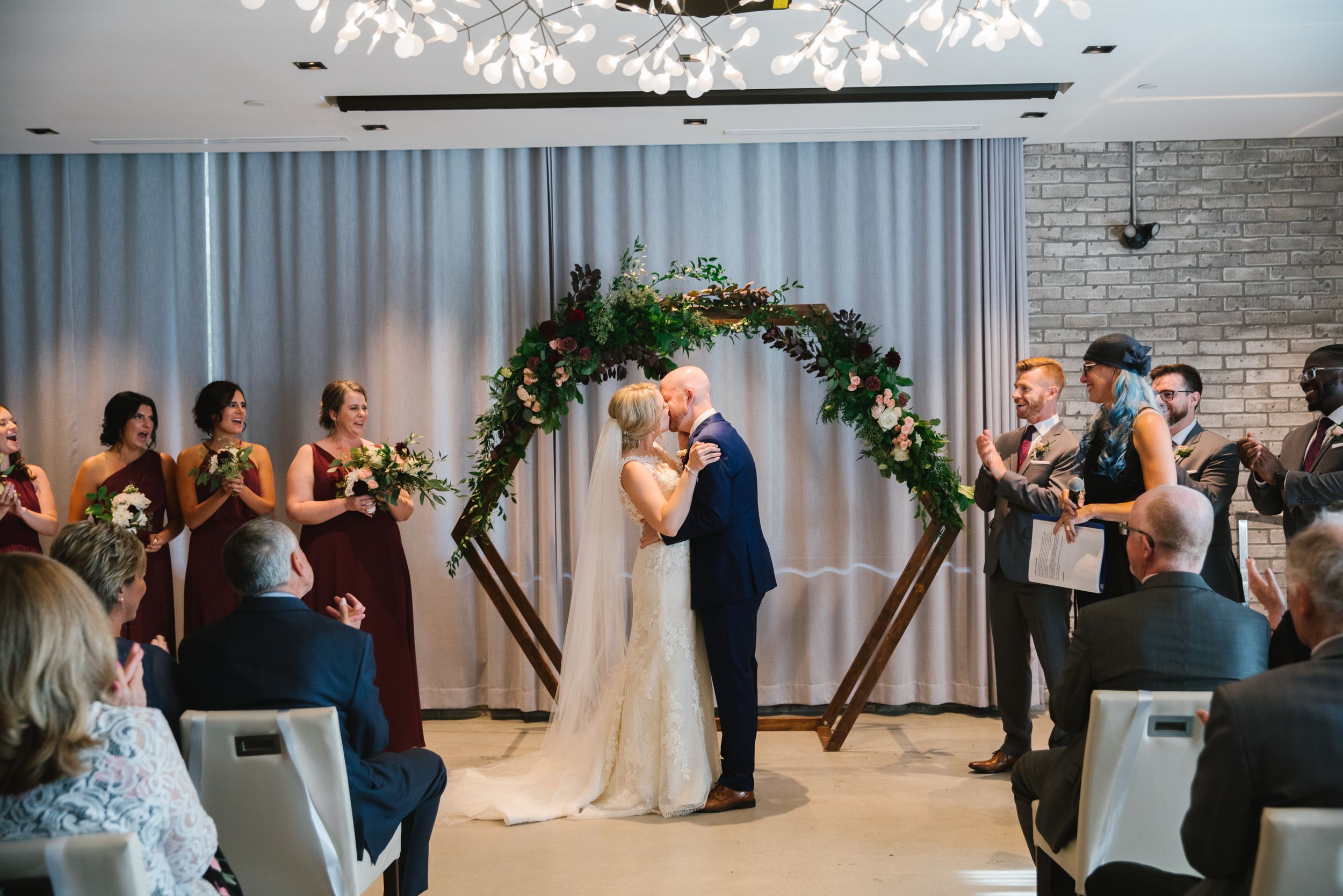bride and groom first kiss hexagonal wedding arch with greenery indoors