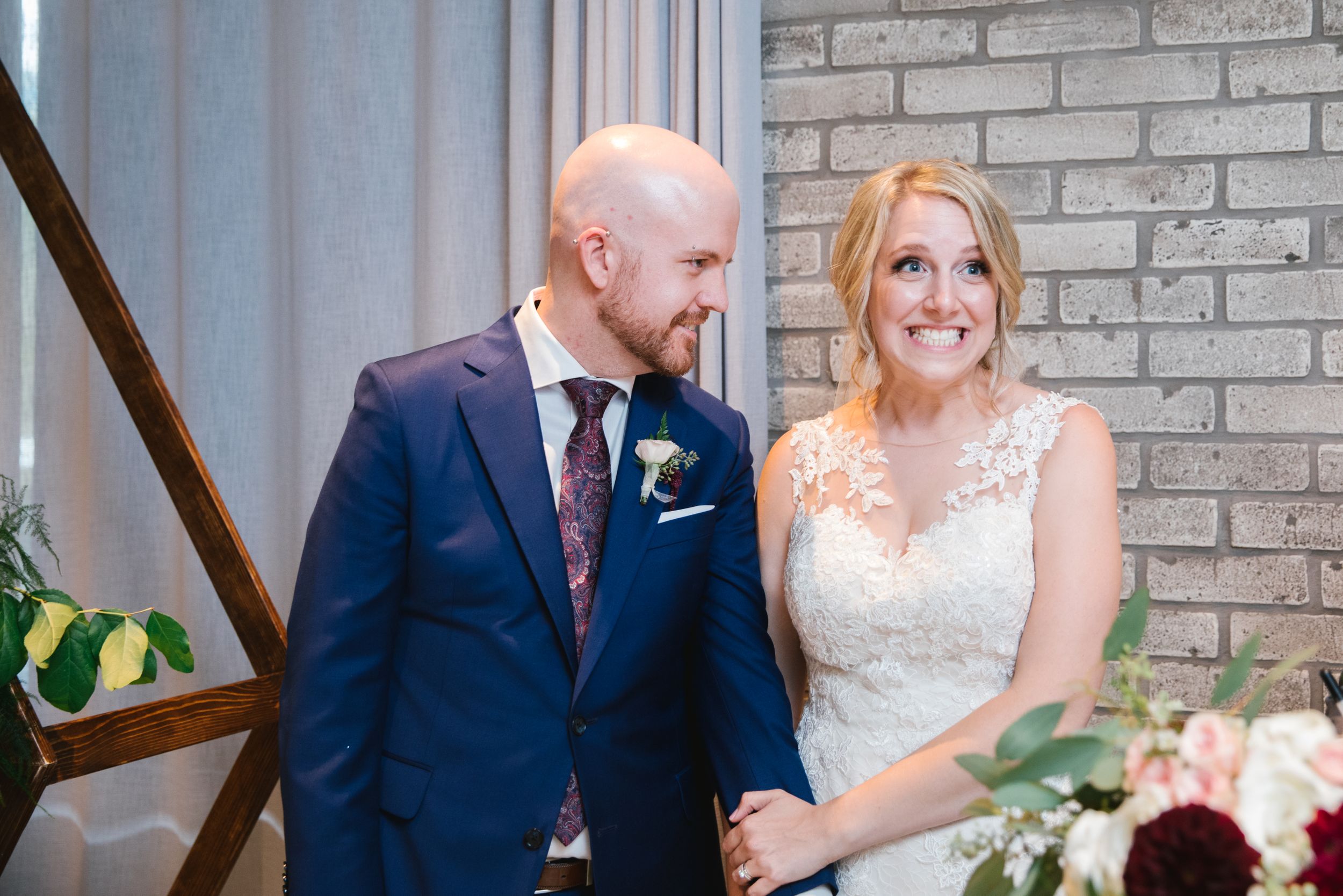 bride pulling a face while groom looks at her and smiles with brick wall behind