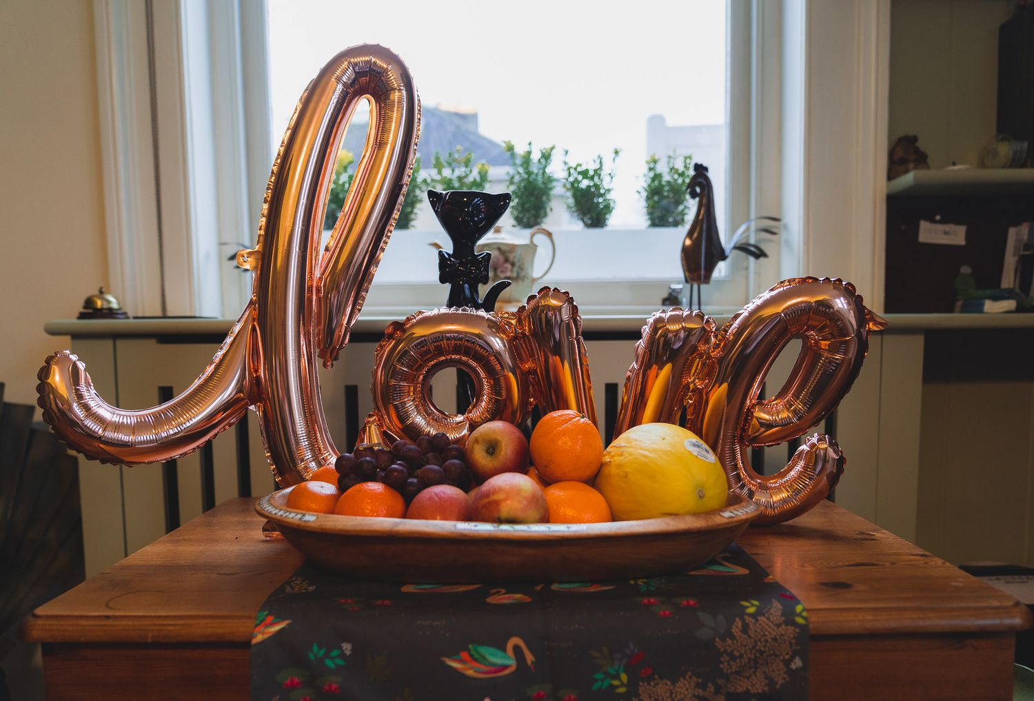 A rose gold love balloon is propped up on a bowl of fruit.