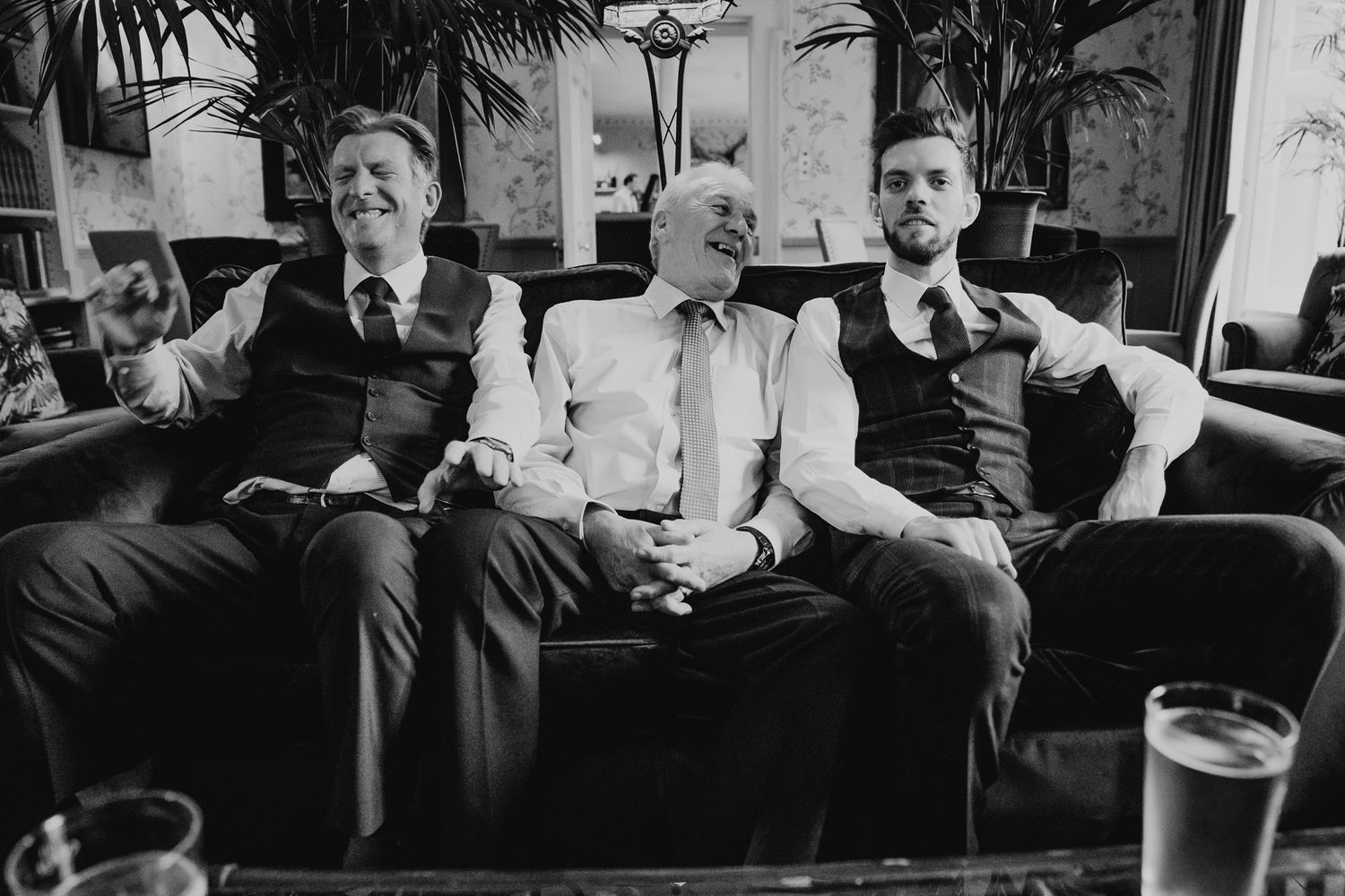 Black and white photo of three men sat laughing on a sofa at a Dorset wedding.