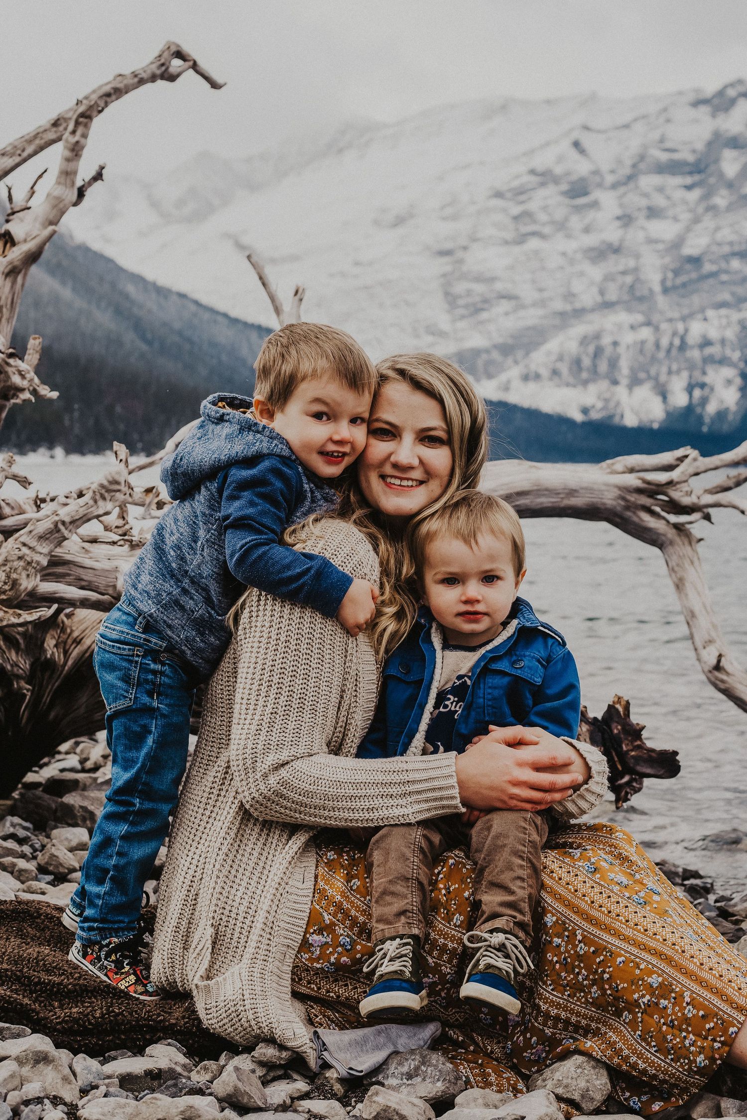 mother holds baby boy while other son hugs her on lake shoreline