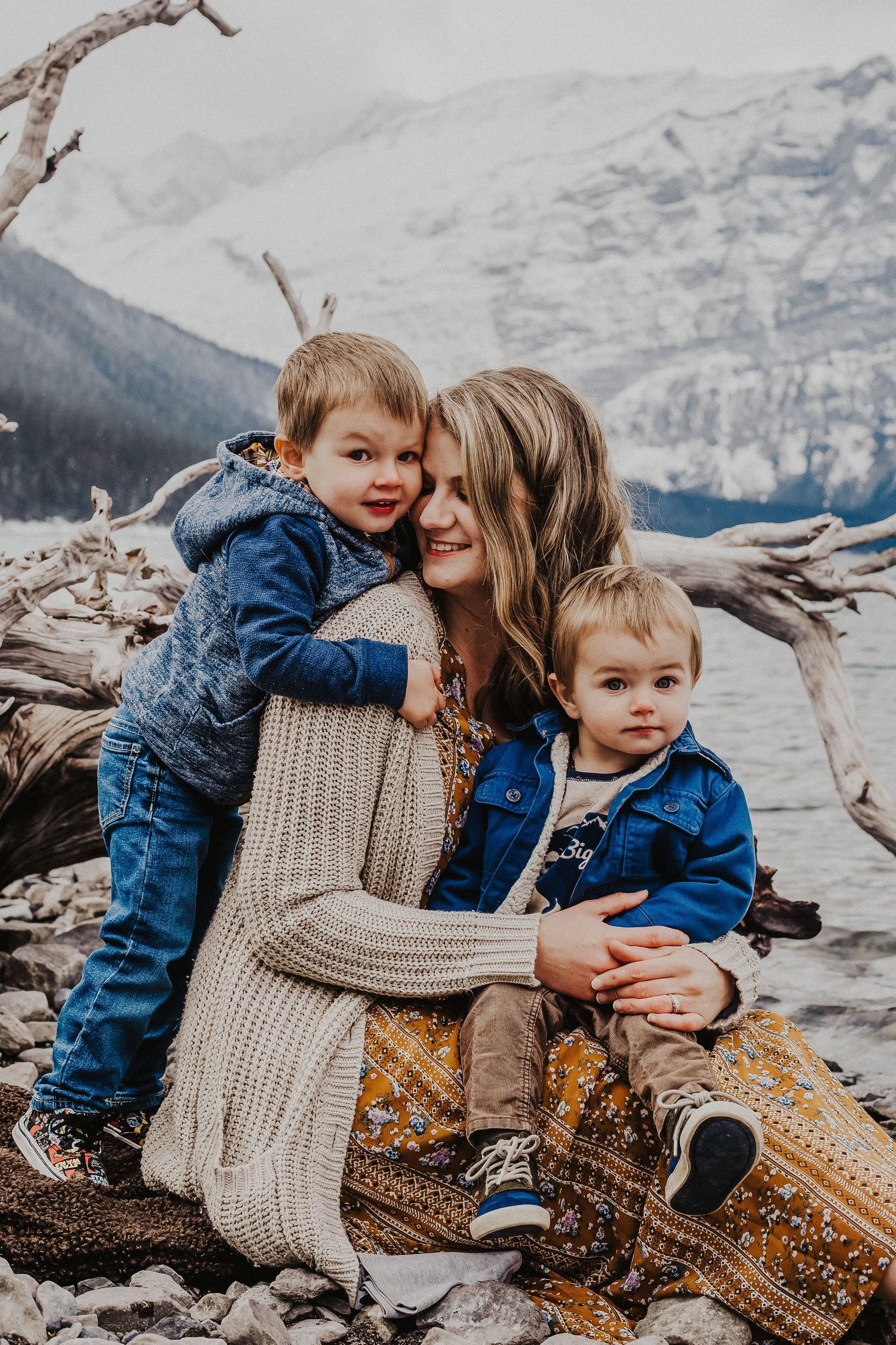 mother holds baby boy while other son hugs her on lake shoreline