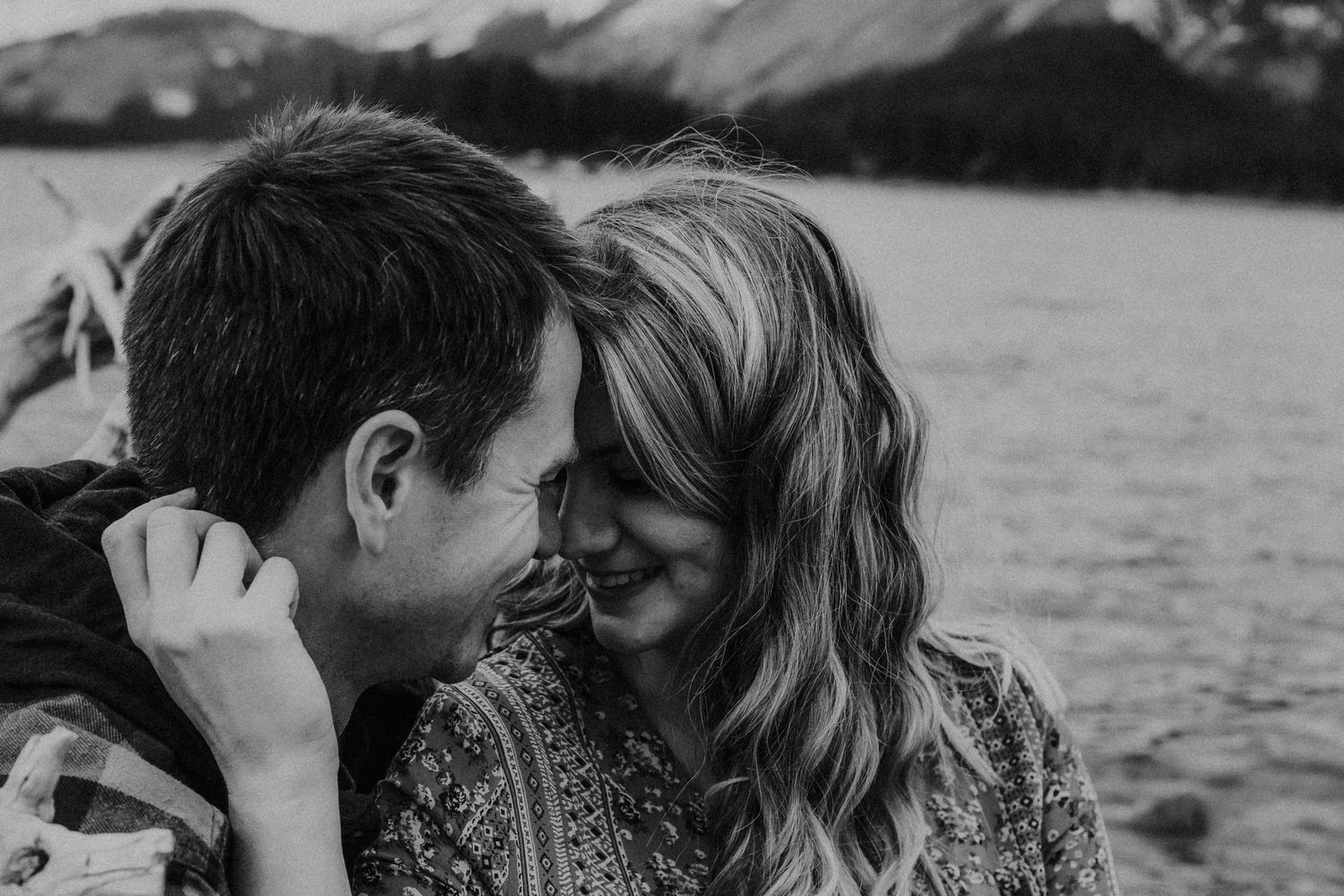 couple smile while face to face sitting on lake shoreline by the mountains