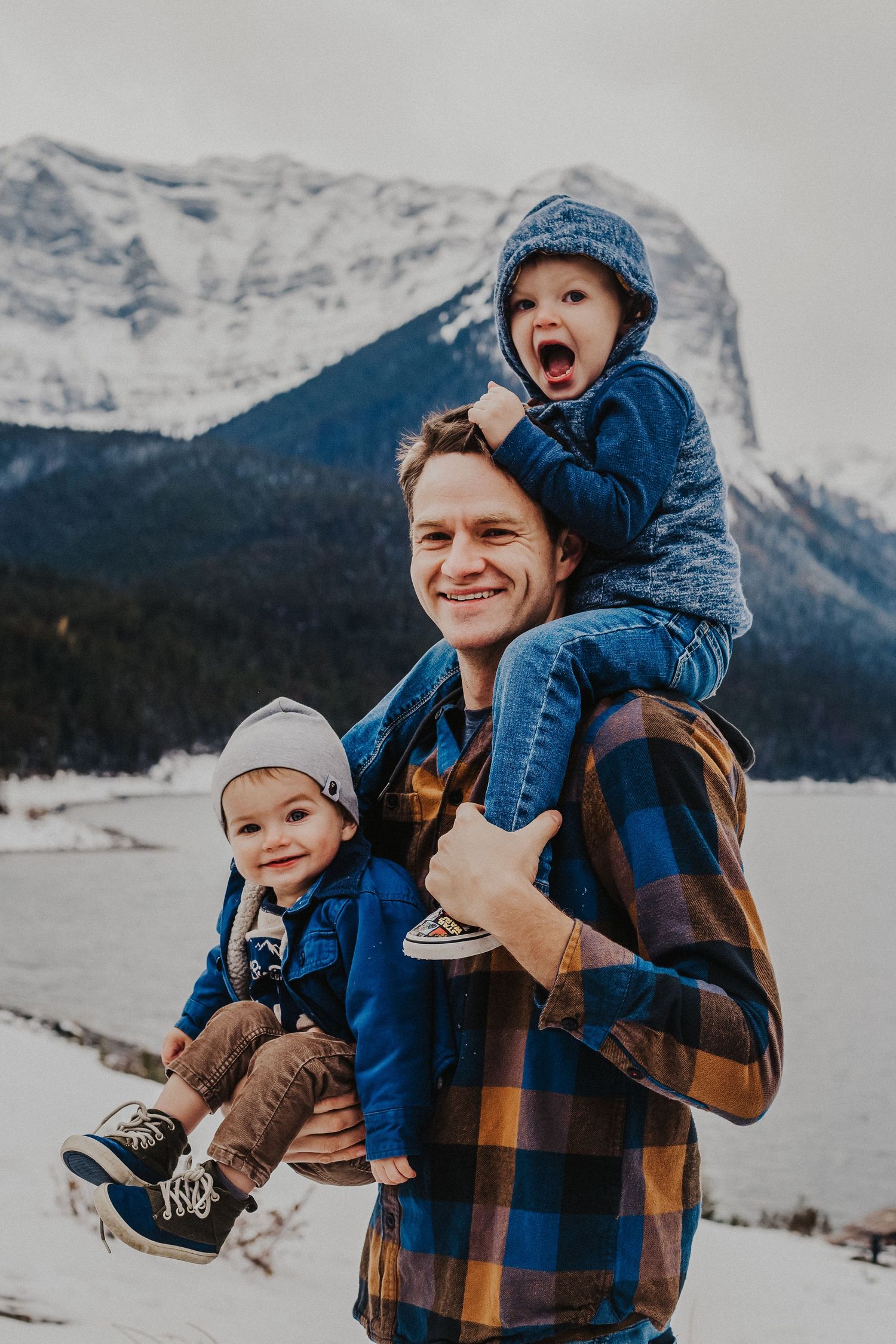 dad holds baby boy and has other son on his shoulders in front of mountains
