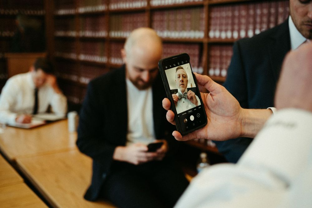 groom-putting-bow-tie-on-with-iphone-camera-wedding-cowles-library-desmoines-iowa-raelyn-ramey-photography