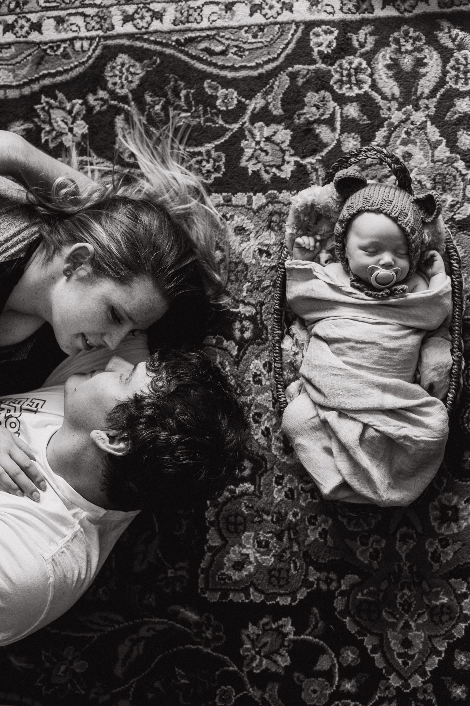 parents look at each other while laying on the floor with newborn baby beside them in a basket