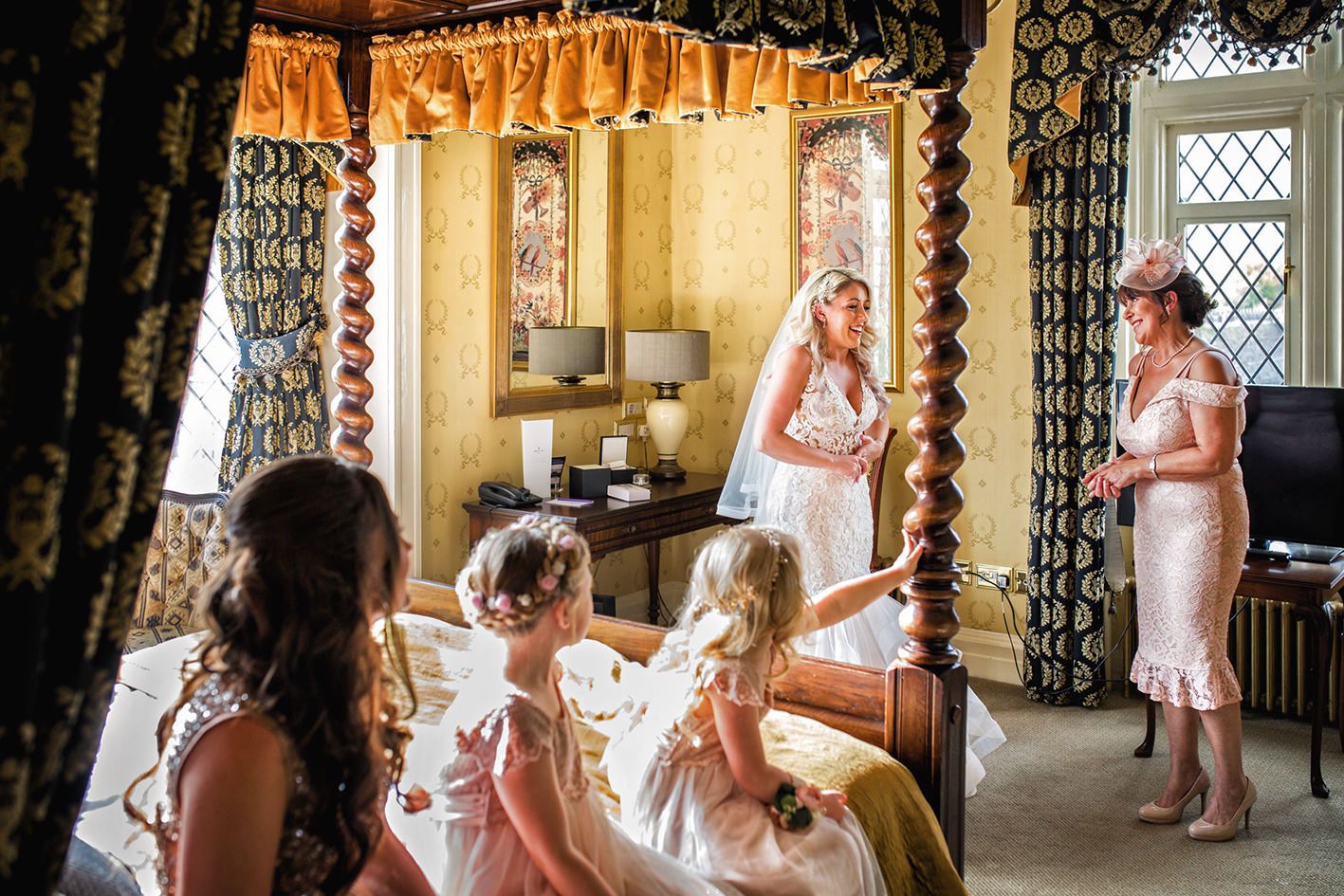 Mother of the bride admires her daughter in a white wedding dress watched by bridesmaids sat on a bed in Crewe Hall