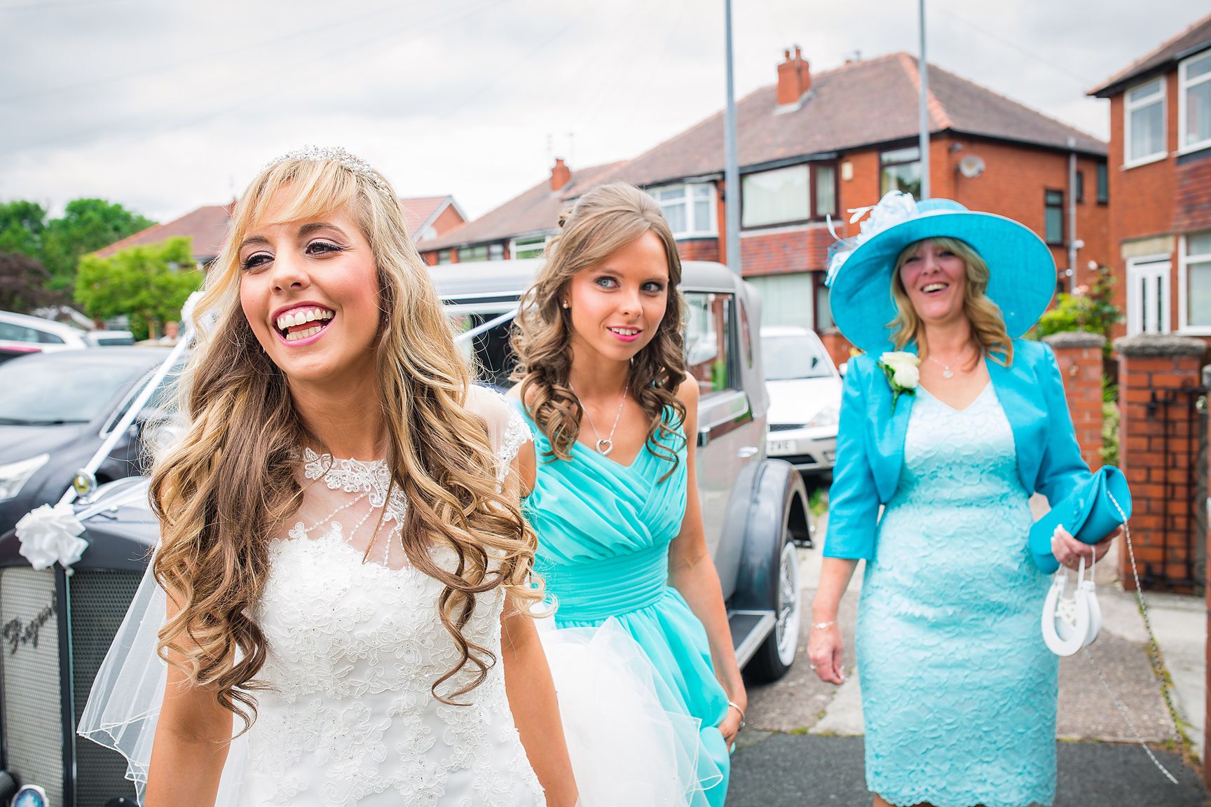 Smiling bride in white wedding dress is watched by her Mother and bridesmaid outside her house