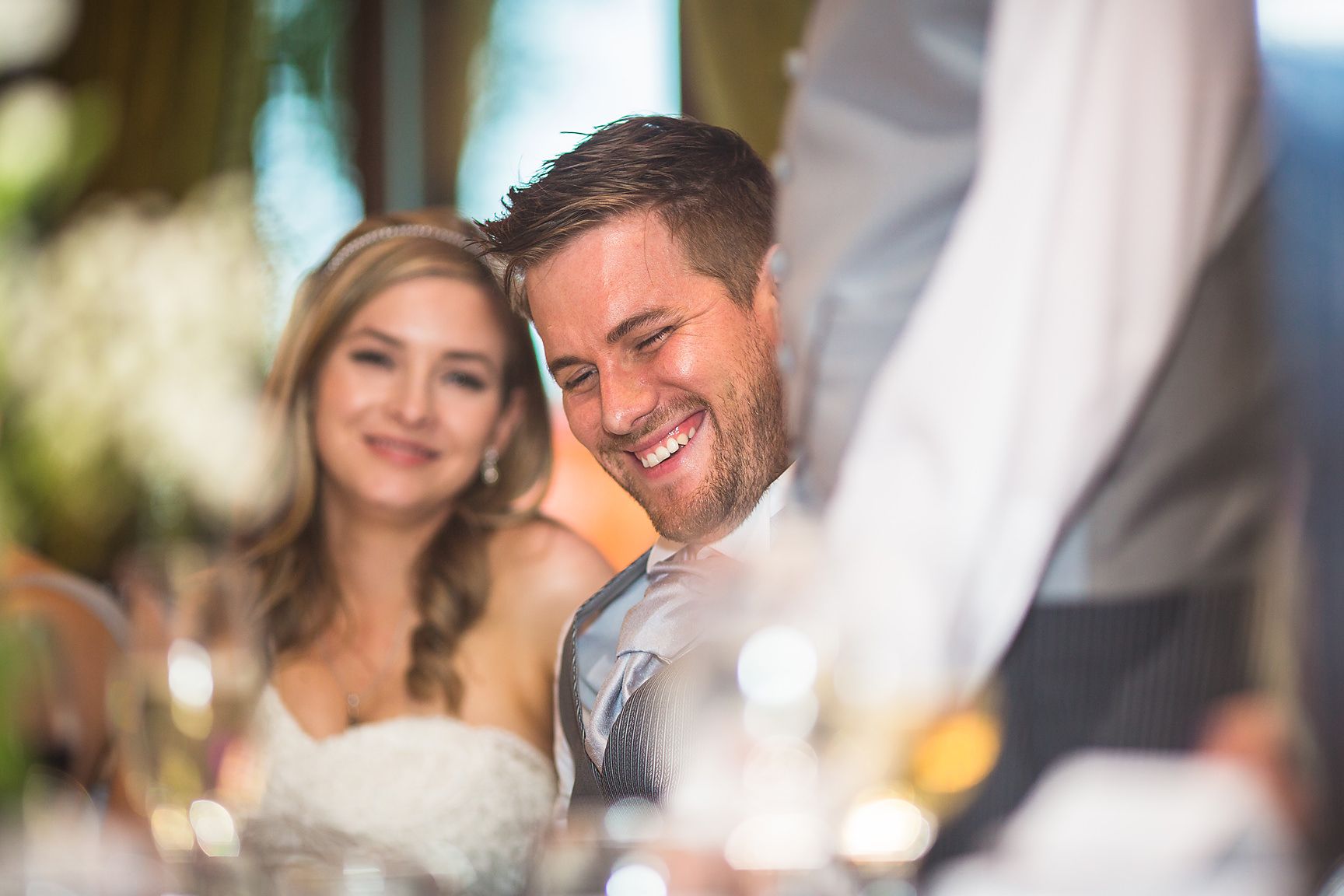 Bride and groom smile at best mans speech at the wedding breafast table