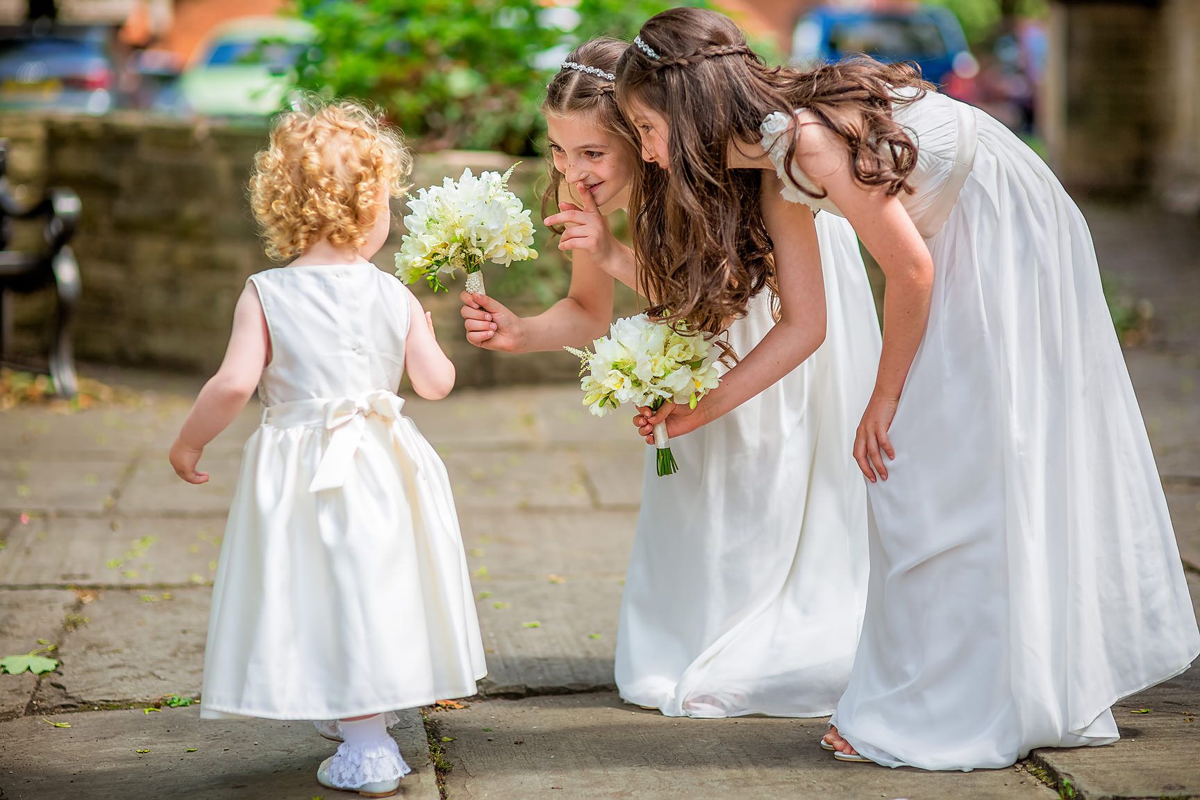 Tiny toddler bridesmaid is greeted by two young bridesmaids all dressed in white outside church