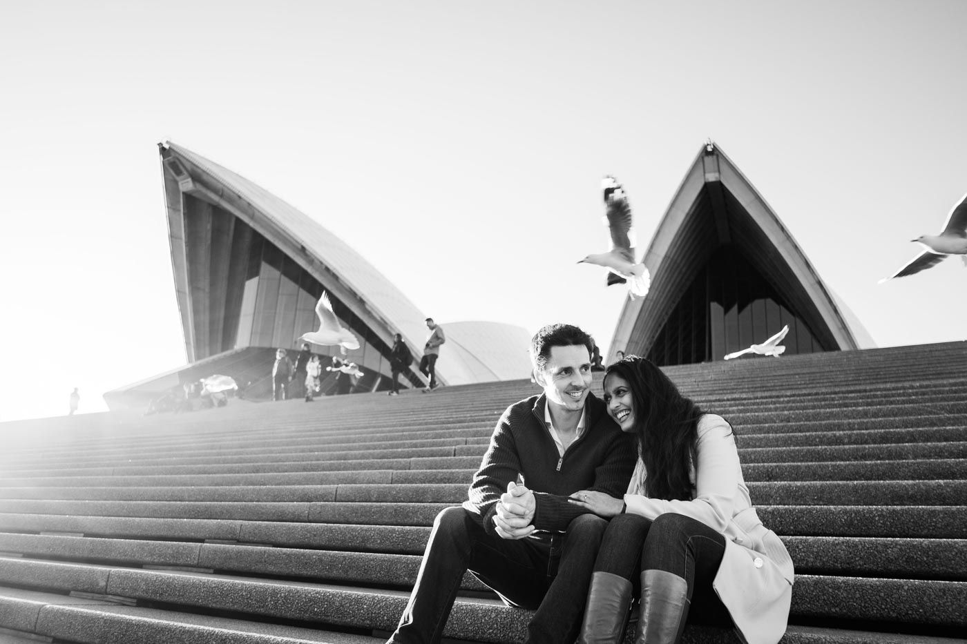 Couple sharing a laugh sitting on the Sydney Opera House steps with seagulls flying overhead