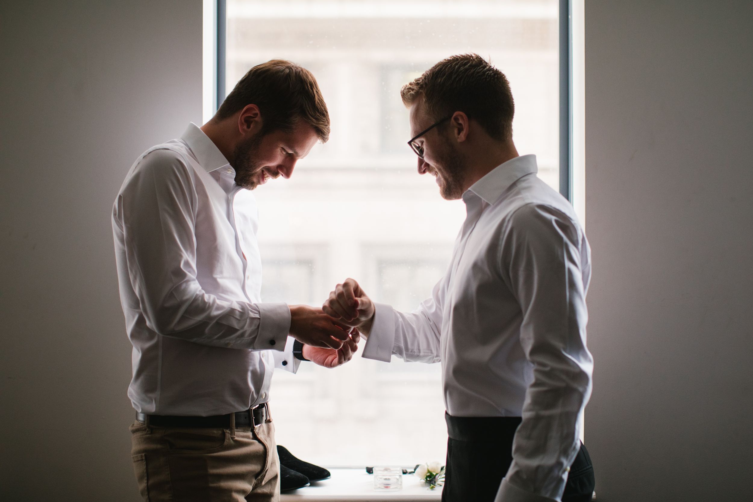 best man helping groom put on cufflinks, backlit by window