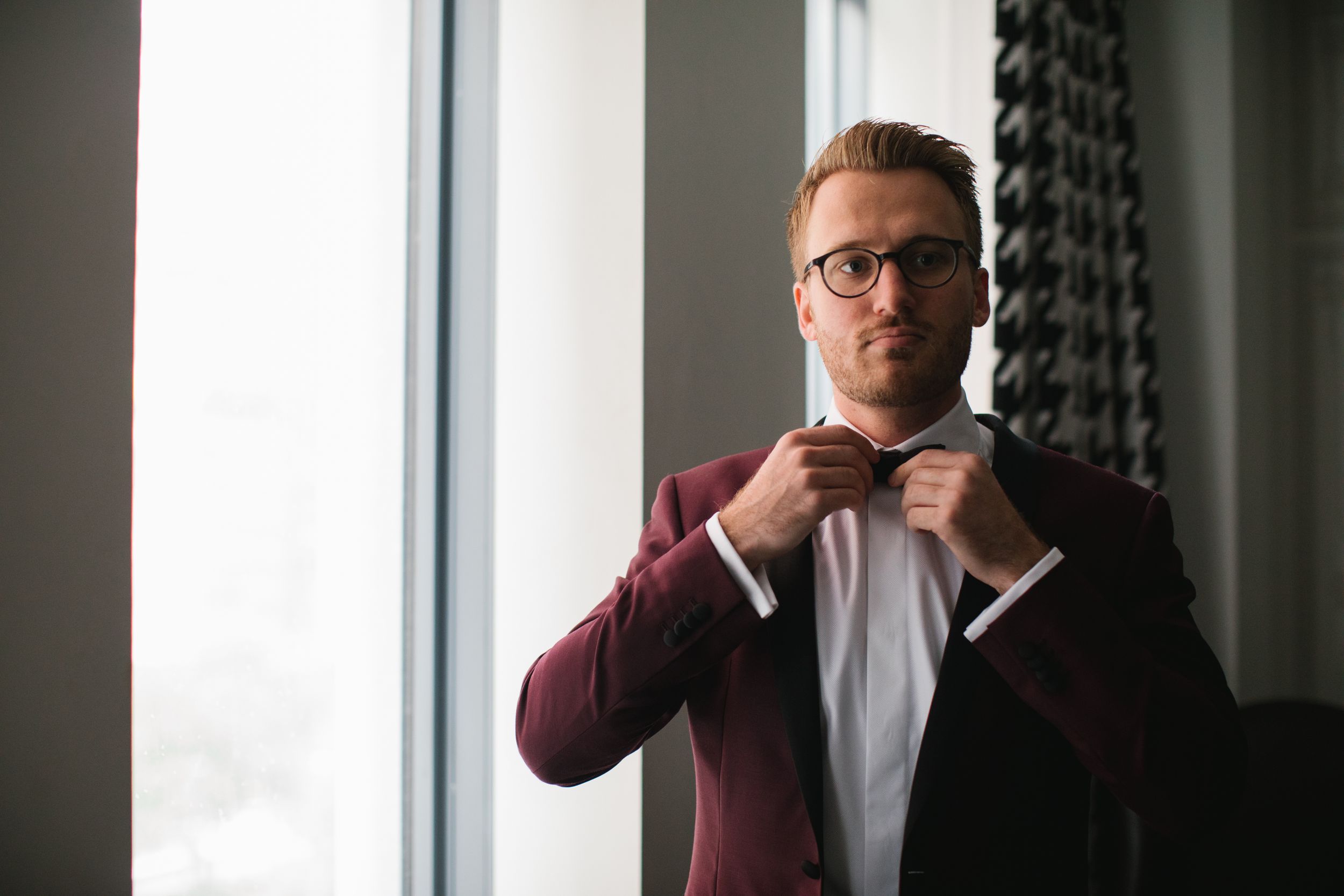 groom putting on bow tie by window