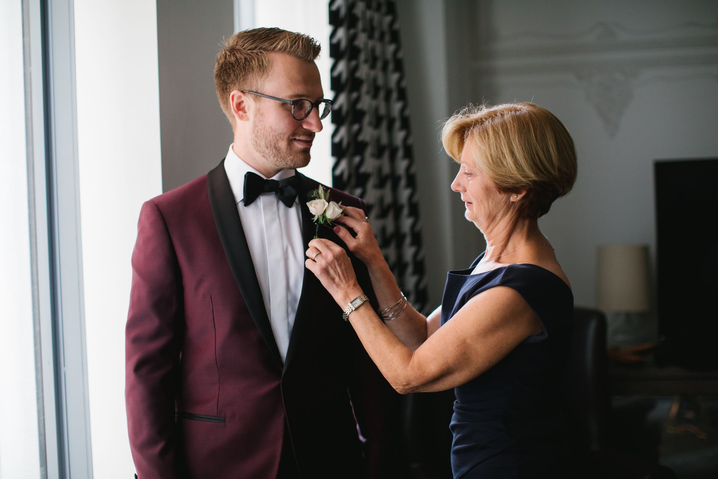 mother of groom helping him put on his boutonniere by window in hotel room