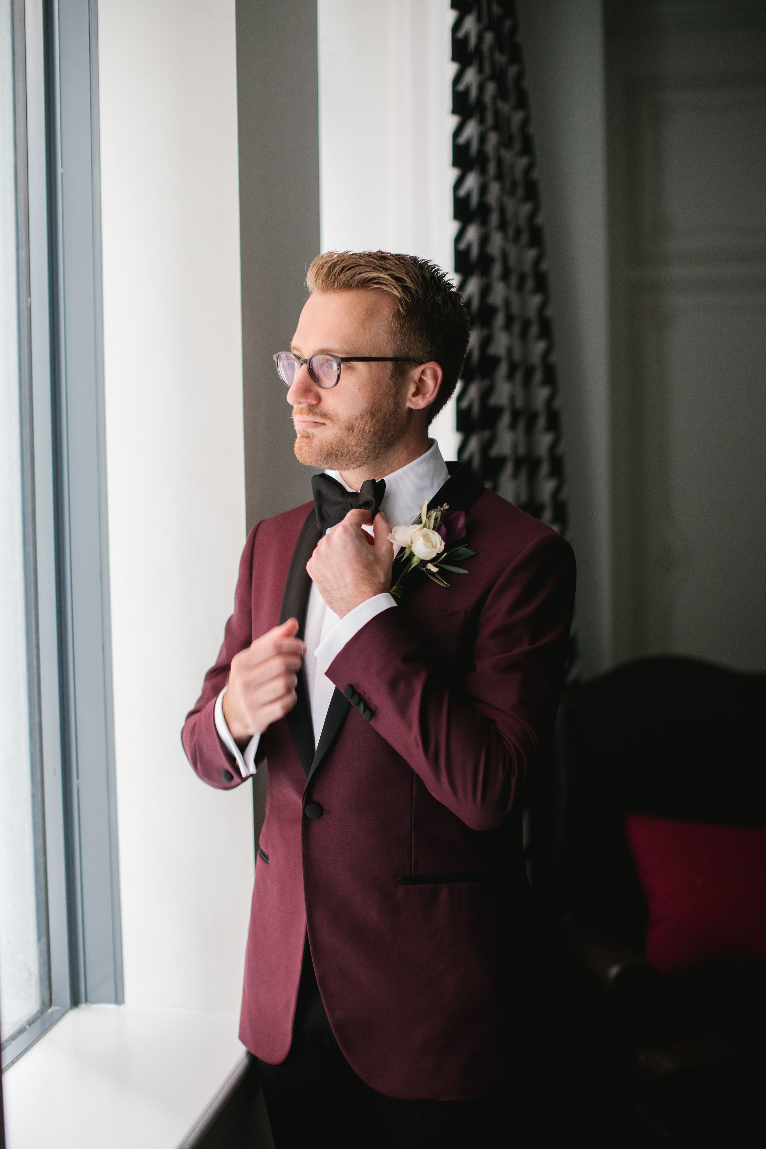 groom portrait by window, wearing a burgundy tuxedo