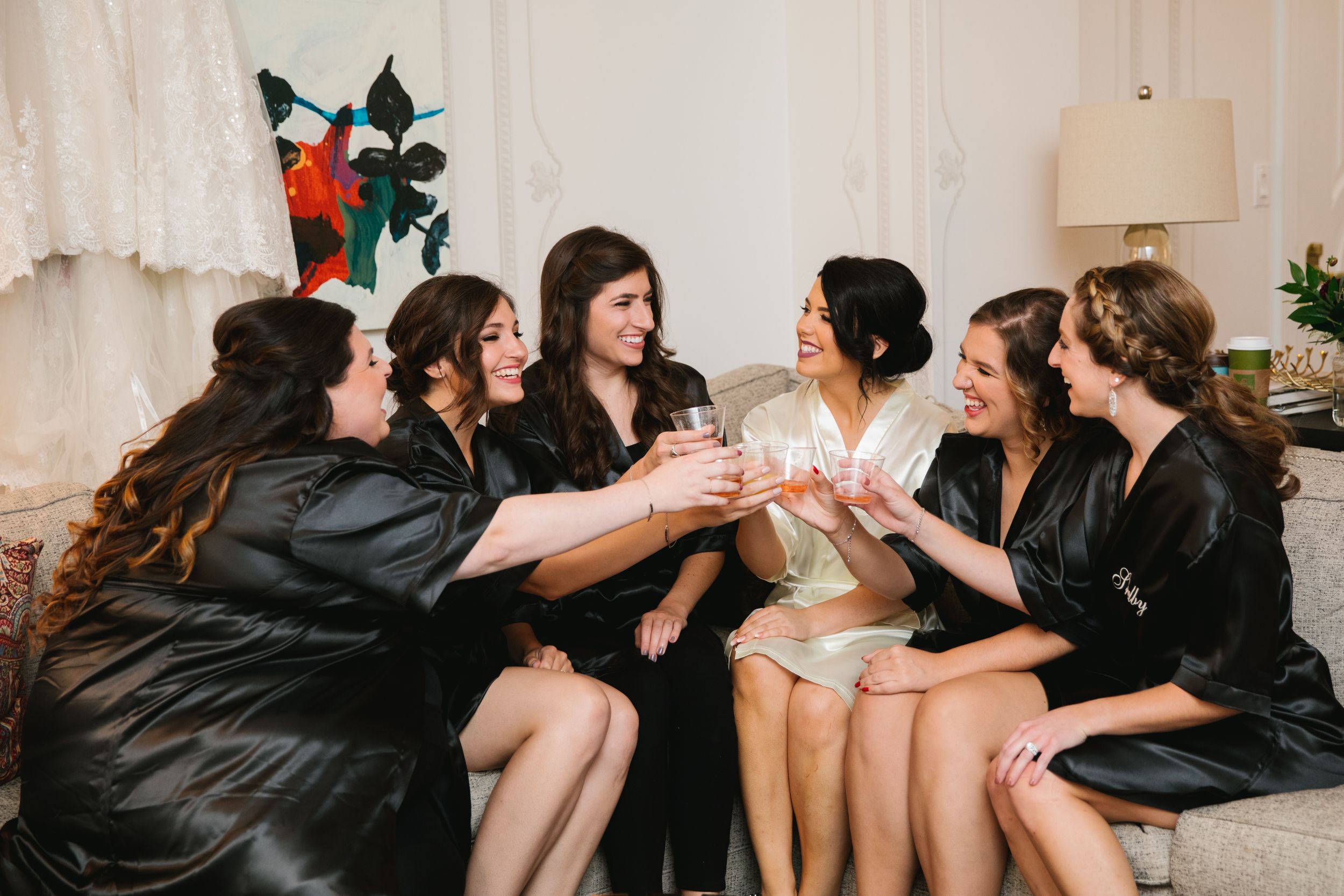 bride with bridesmaids in robes sitting on sofa and cheersing with champagne