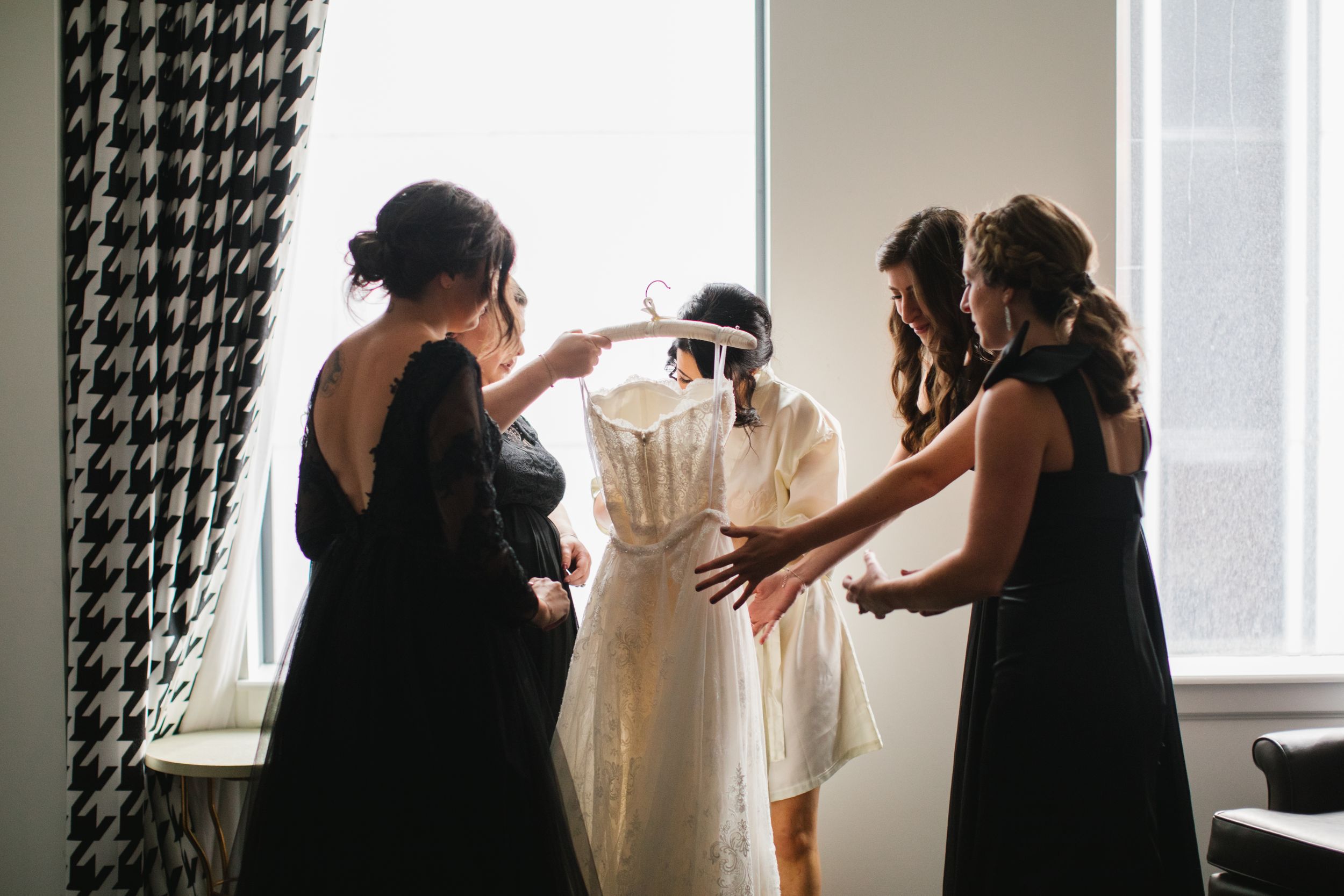 bride with bridesmaids putting on wedding dress in front of window