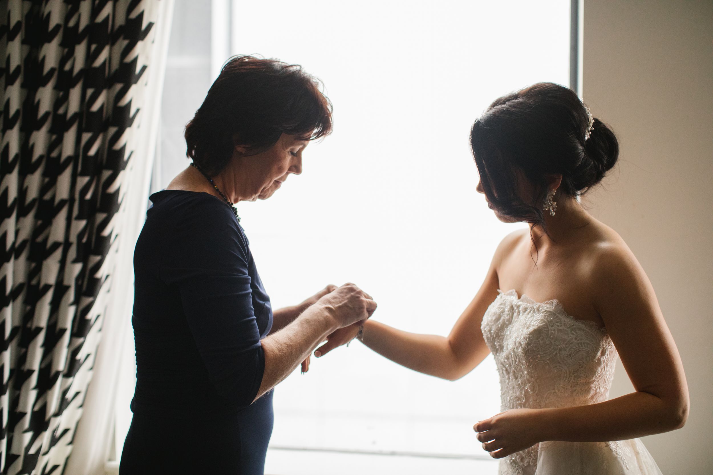 bride and mother of bride standing in front of window putting on jewellery