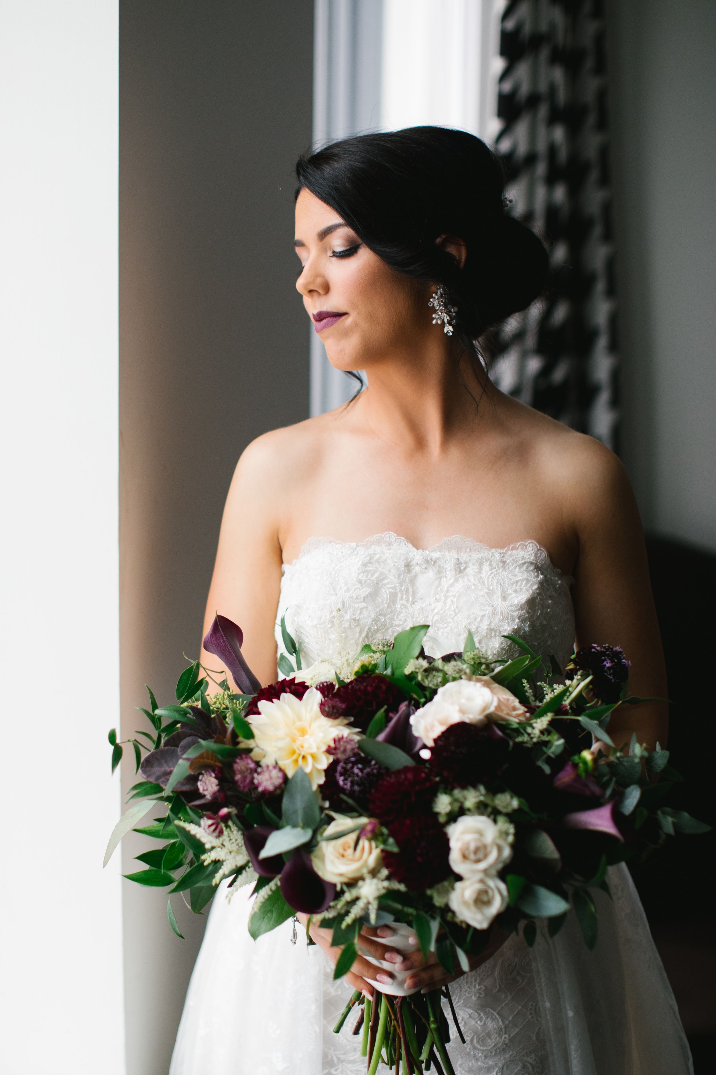 bridal portrait next to window holding bouquet with burgundy and white florals for wedding