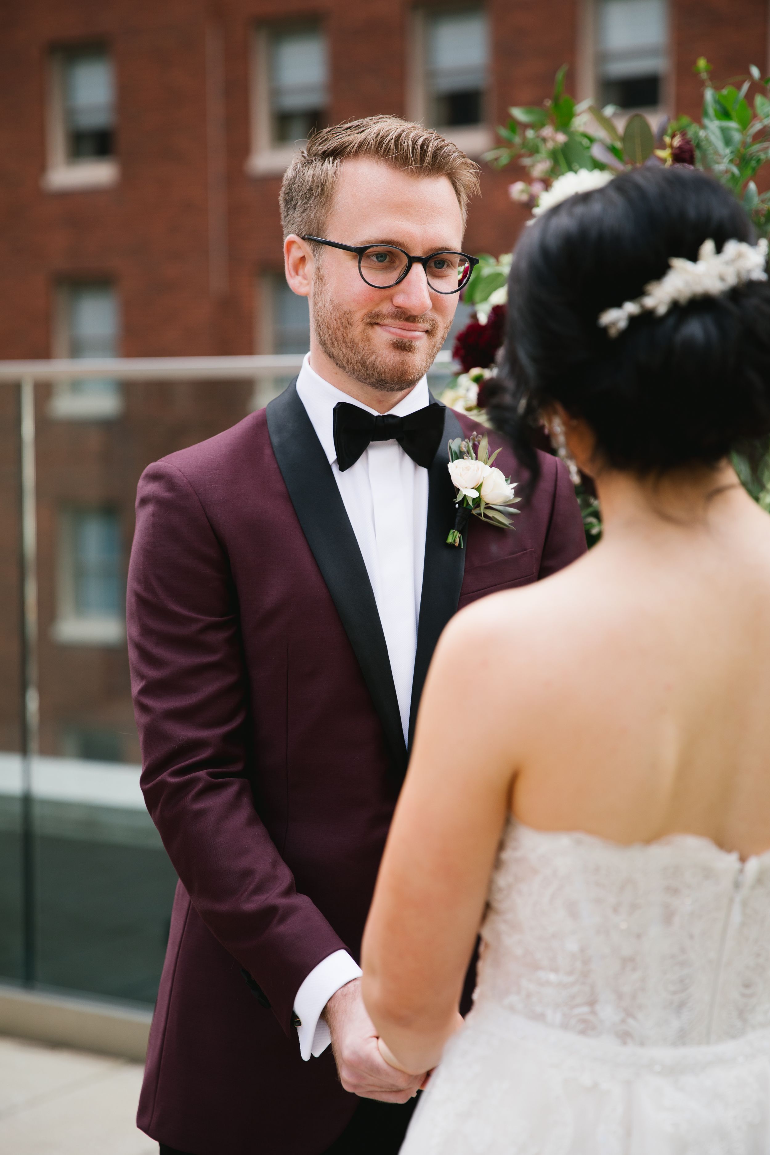 groom wearing a burgundy tuxedo for wedding holding bride's hands and looking into her eyes during rooftop ceremony