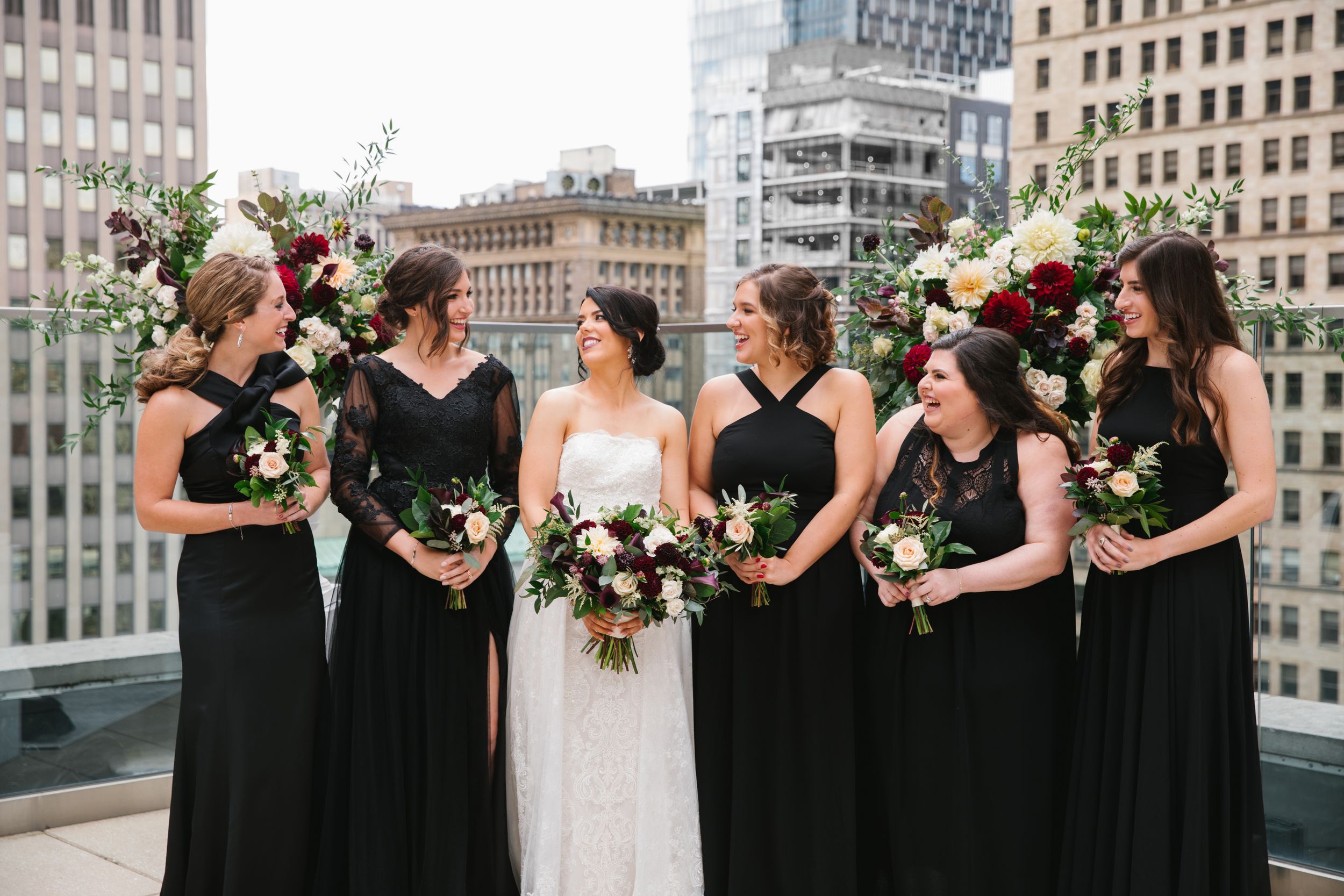 bride and bridesmaids in black dresses holding burgundy bouquets and laughing with city scape in the background