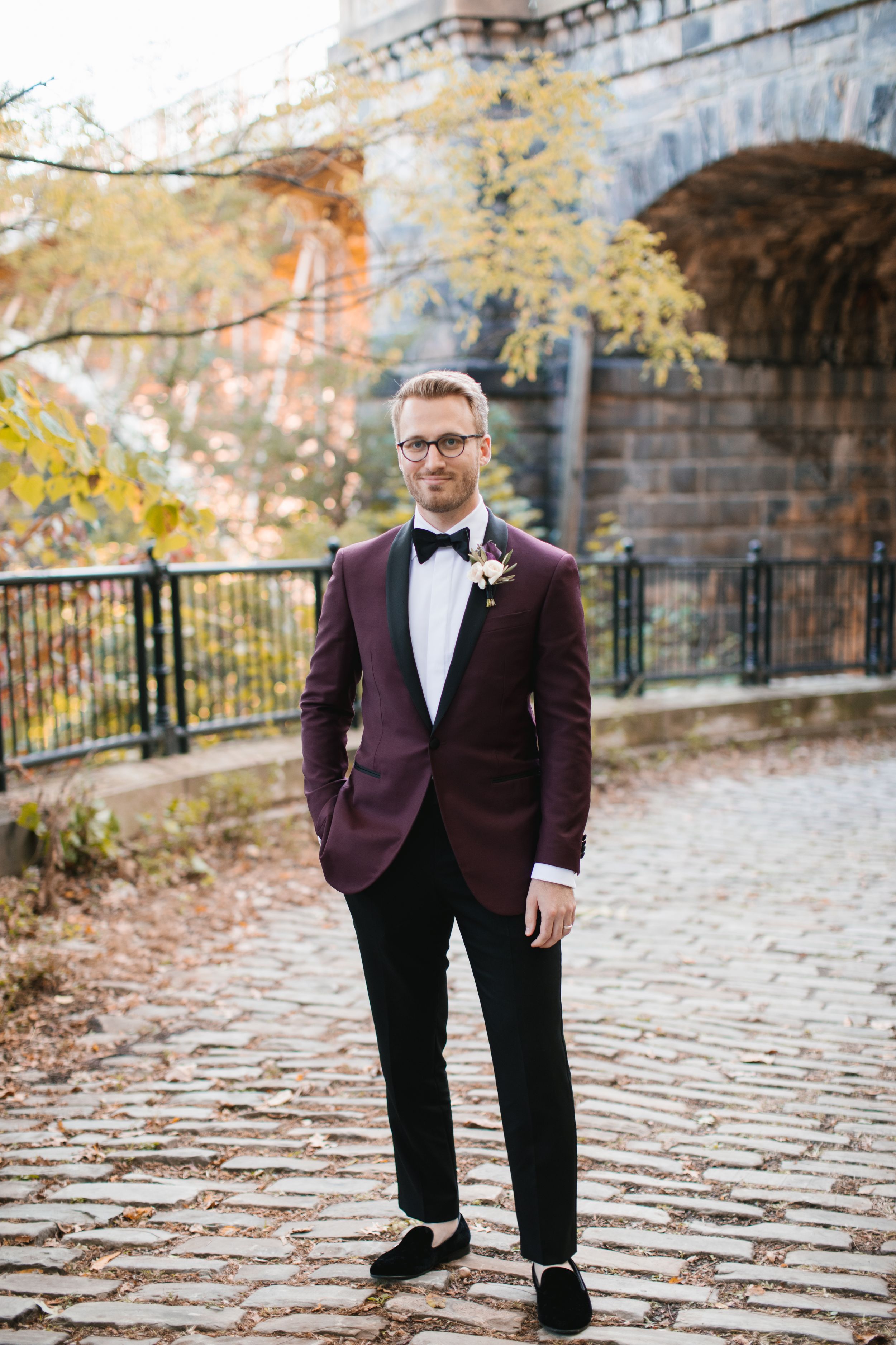 groom portrait wearing burgundy and black tuxedo on cobble stone path in park during fall