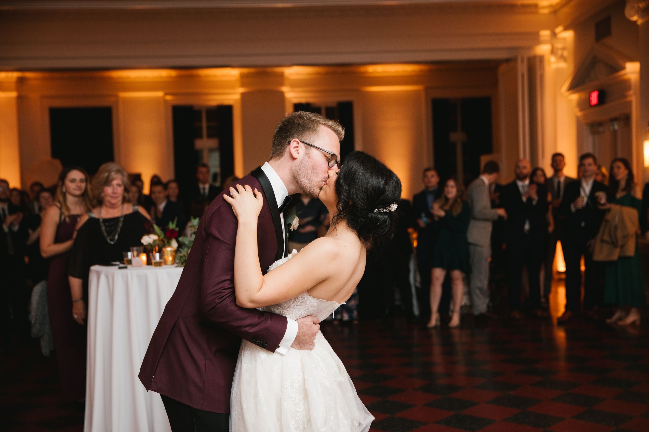 bride and groom kiss during first dance with friends and family behind them