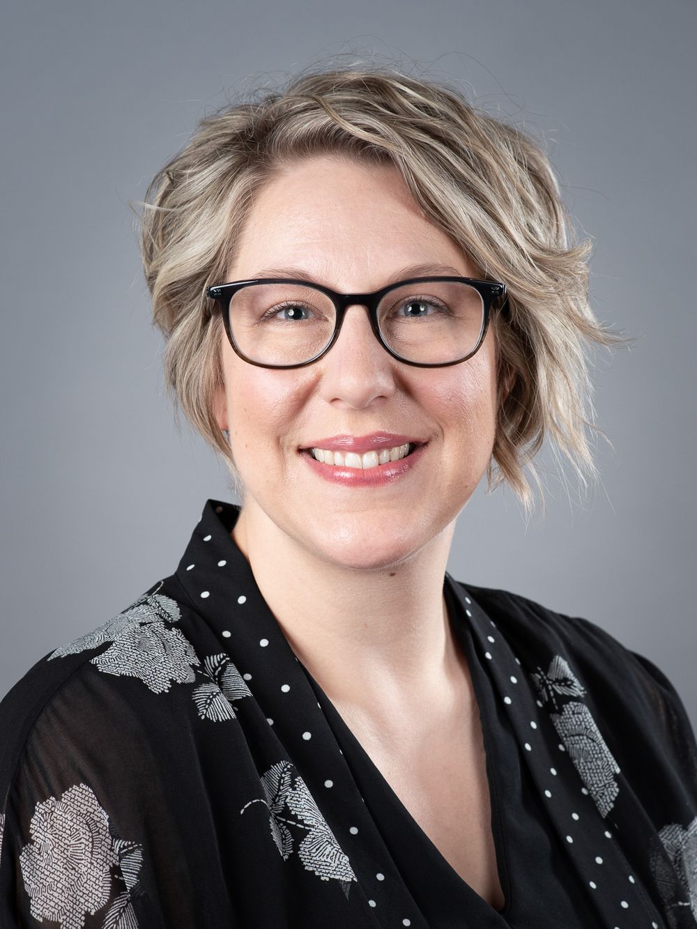 Picture with gray background of attractive professional-looking woman with blonde-streaked hair and black eyeglasses.