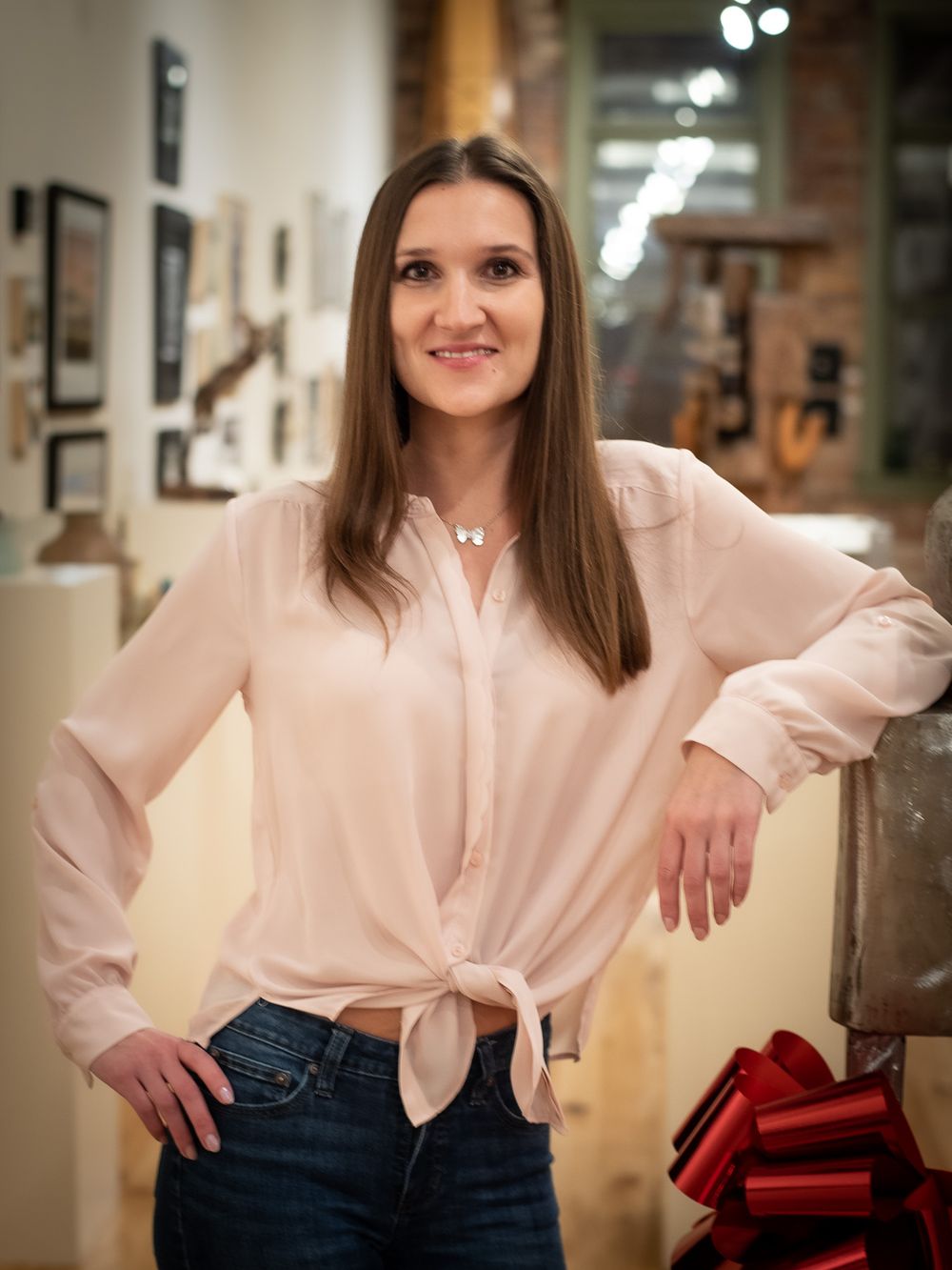 Long-haired woman in a long-sleeve blouse standing with her elbow on a metal sculpture in an art gallery.
