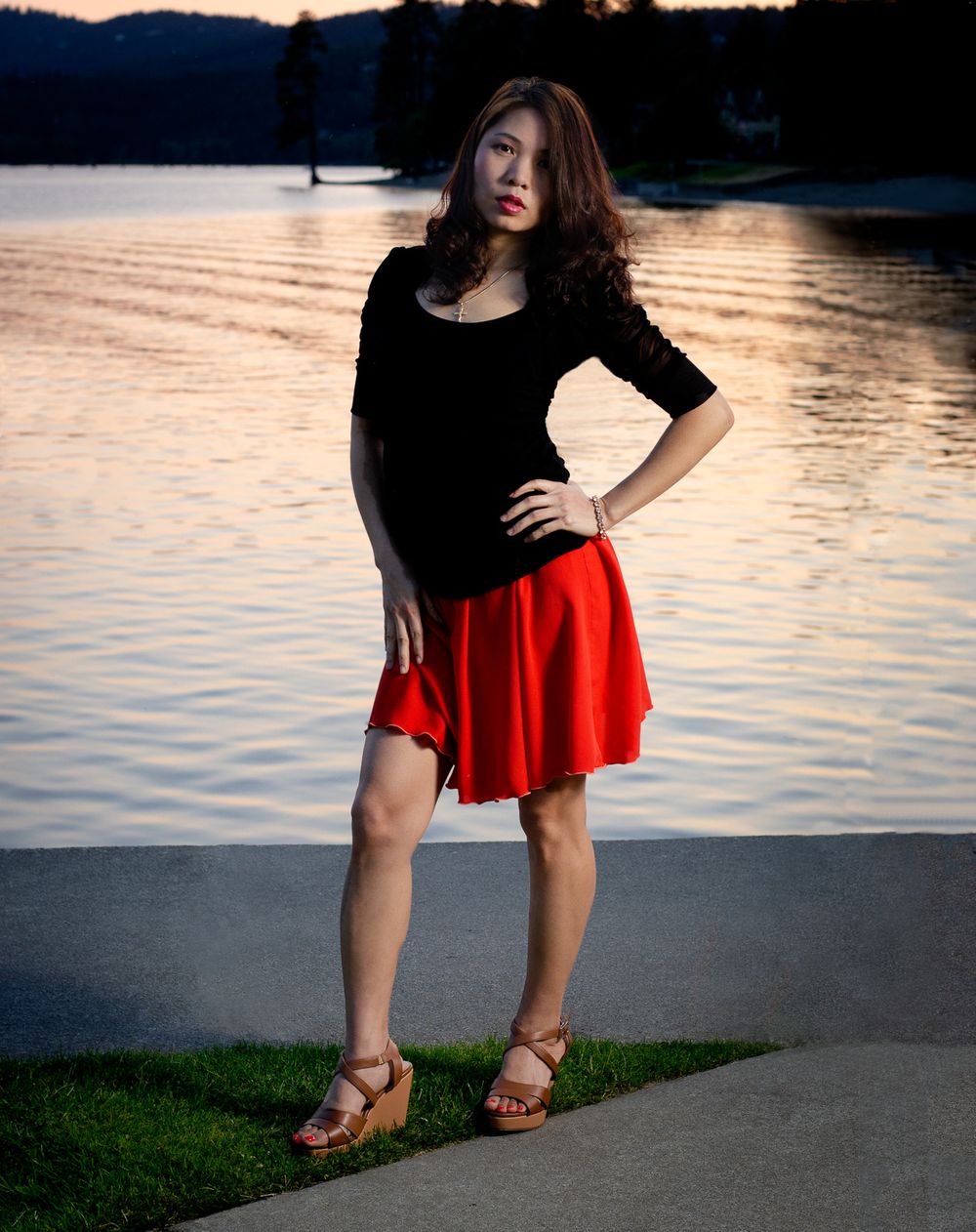Asian woman in black top and red skirt posing in front of a lake at sunset.