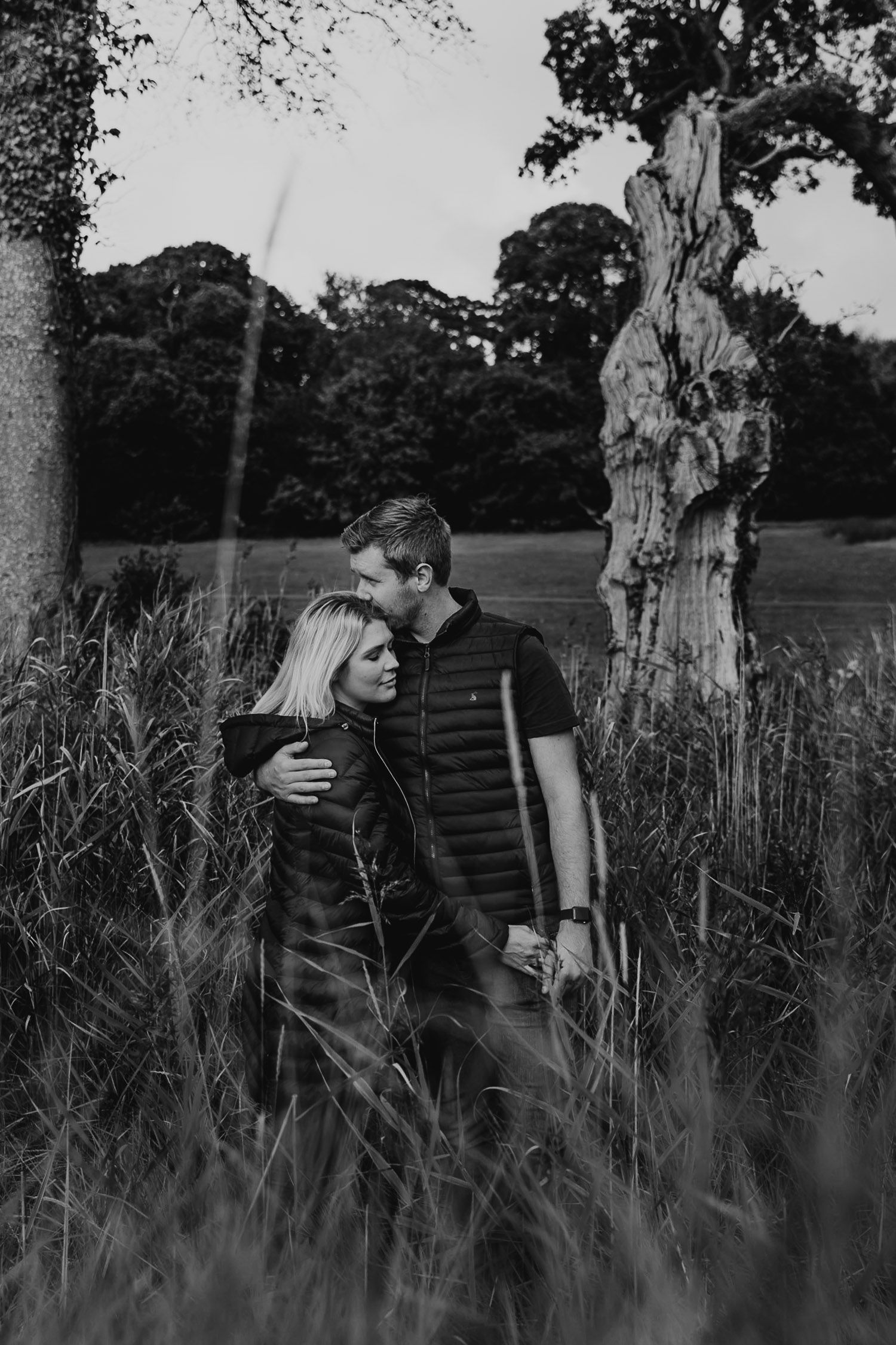 Black and white image. A man wraps his arm around his fiance and kisses her on the forehead. Katherine and her Camera