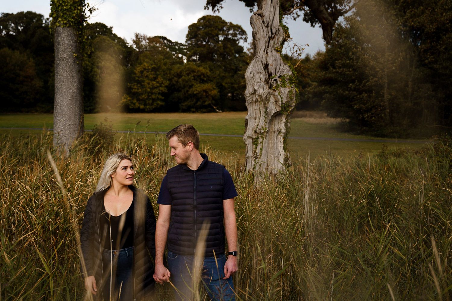 Engaged couple stand in long grass holding hands and looking at each other. Katherine and her Camera.