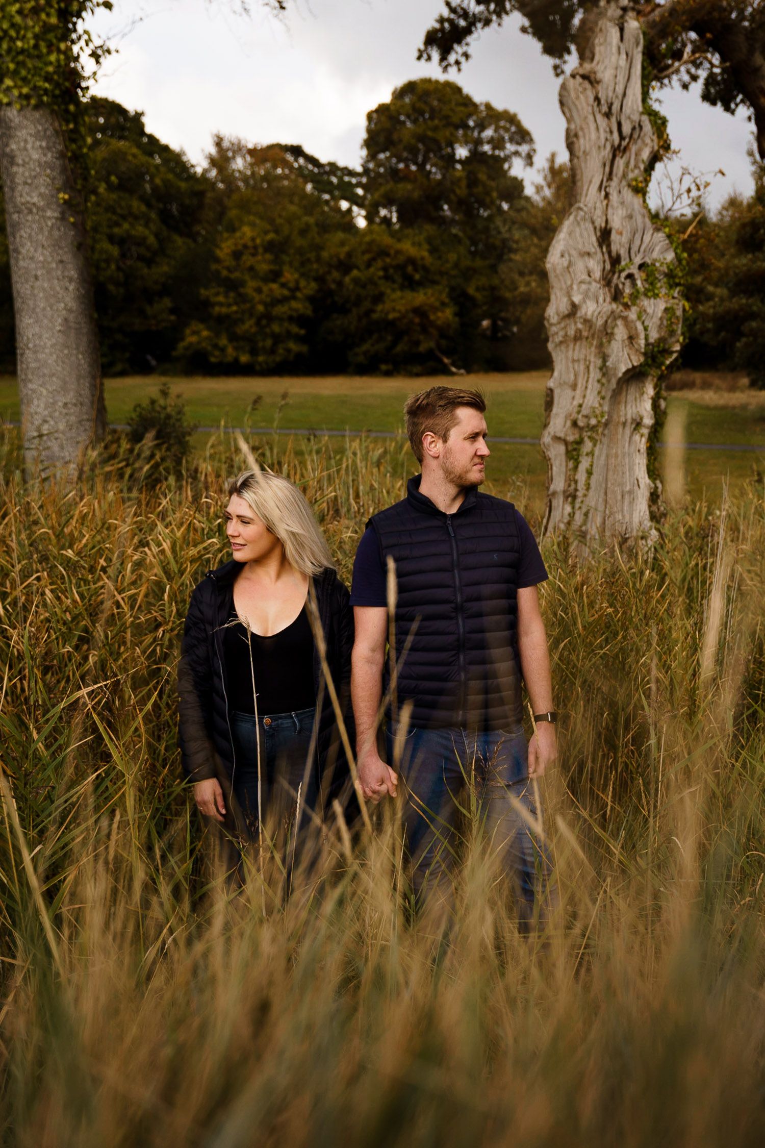 Couple stand in tall grass holding hands and looking in opposite directions. Katherine and her Camera