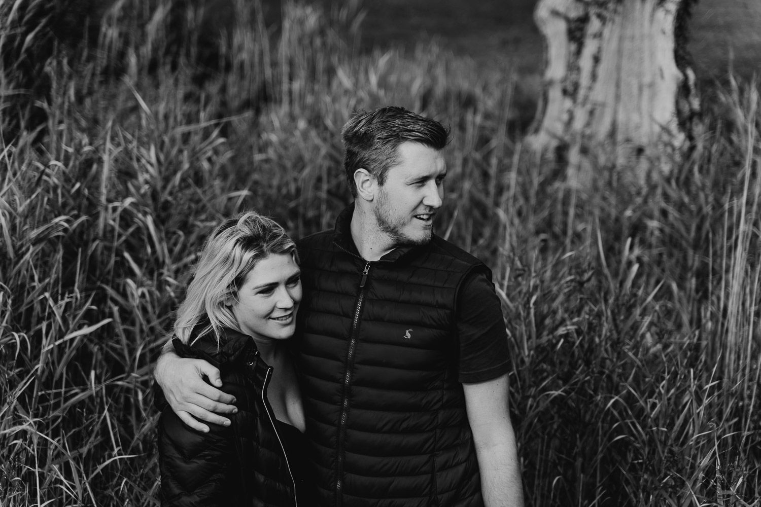 Black and white image, couple cuddle up together in long grass. Katherine and her Camera engagement photoshoot.