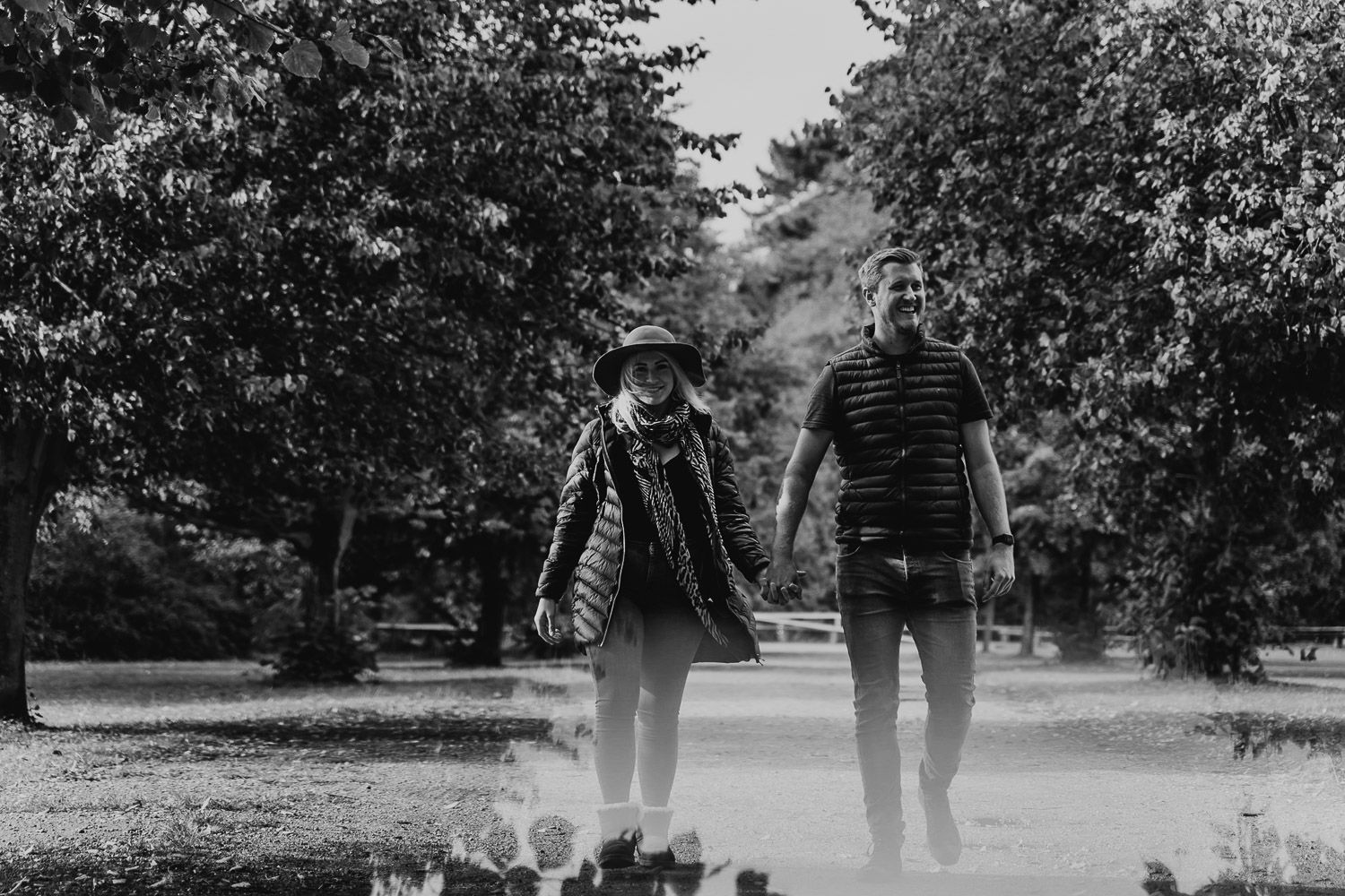 Black and white image of a couple walking in a park with a reflection of the trees at their feet.