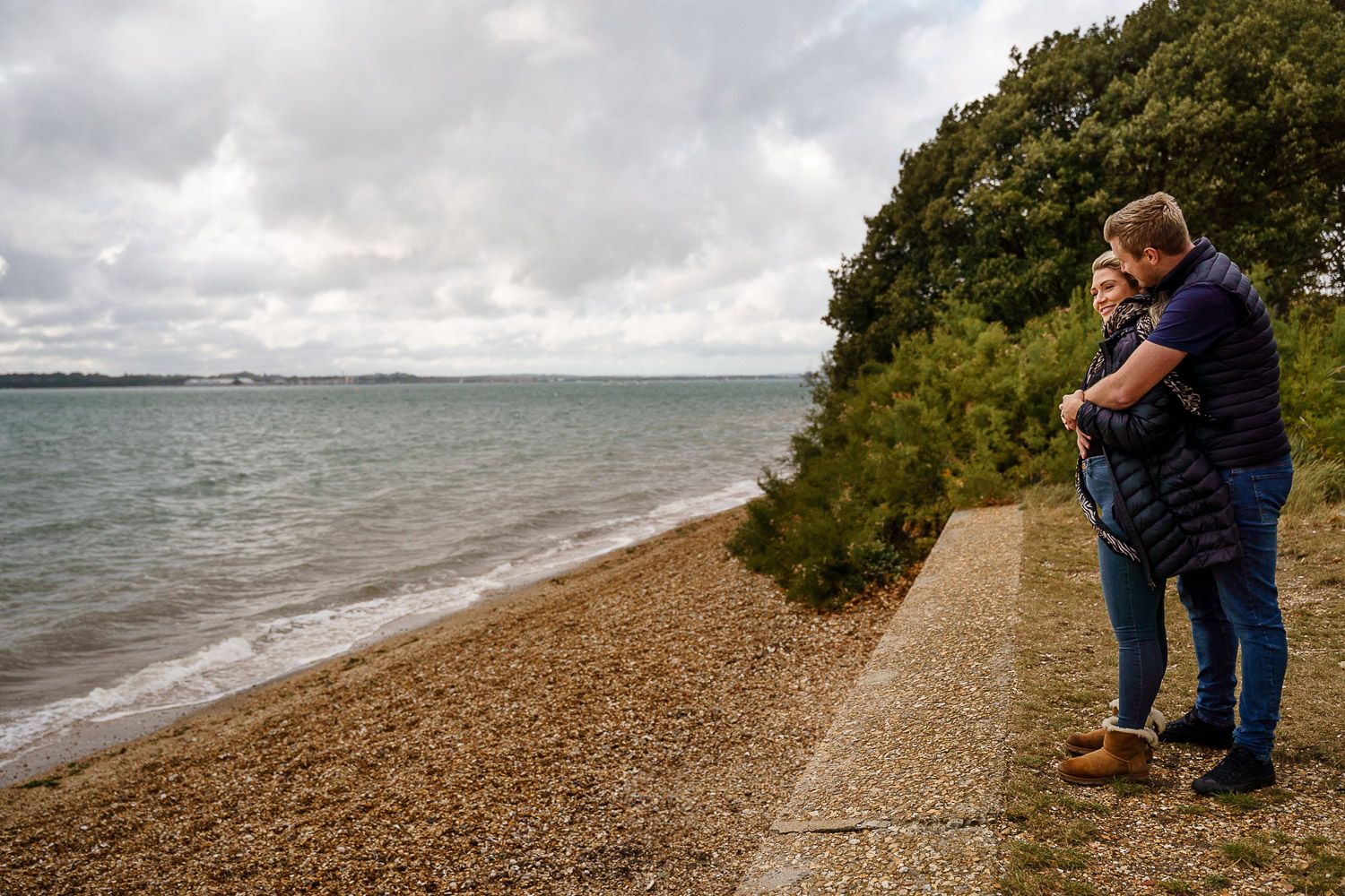 Couple cuddle on a shingle beach and look out to the sea. Katherine and her Camera engagement photoshoot.