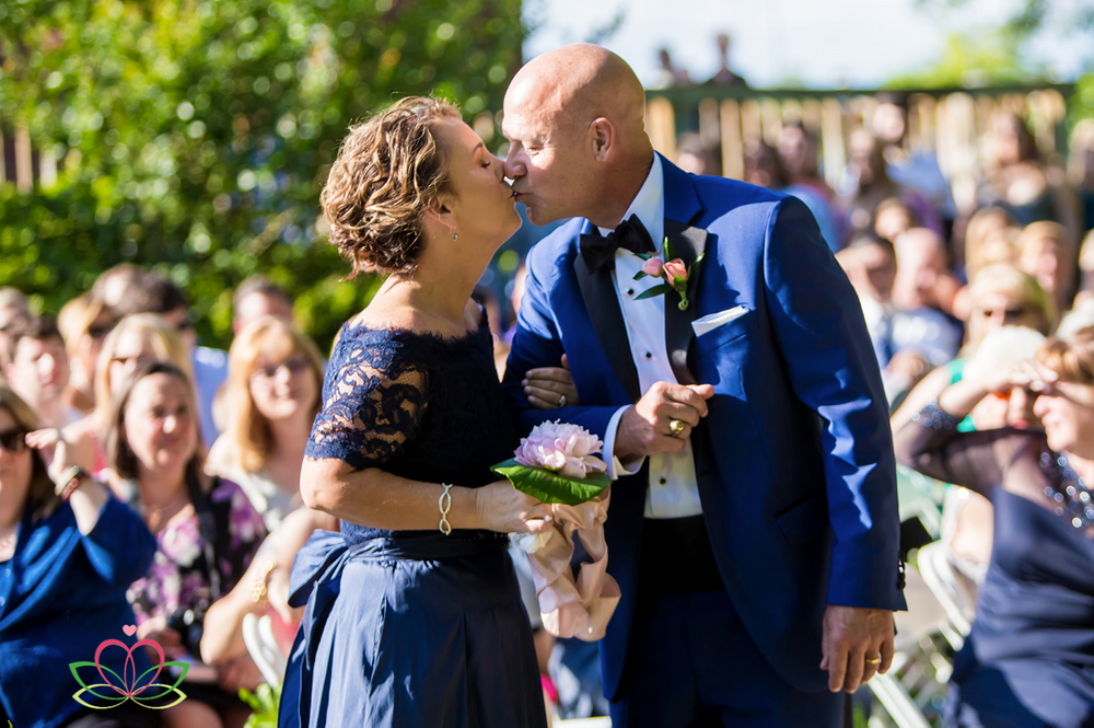 The father of the bride kisses the mother of the bride after walking the bride during a wedding ceremony at Senate's End
