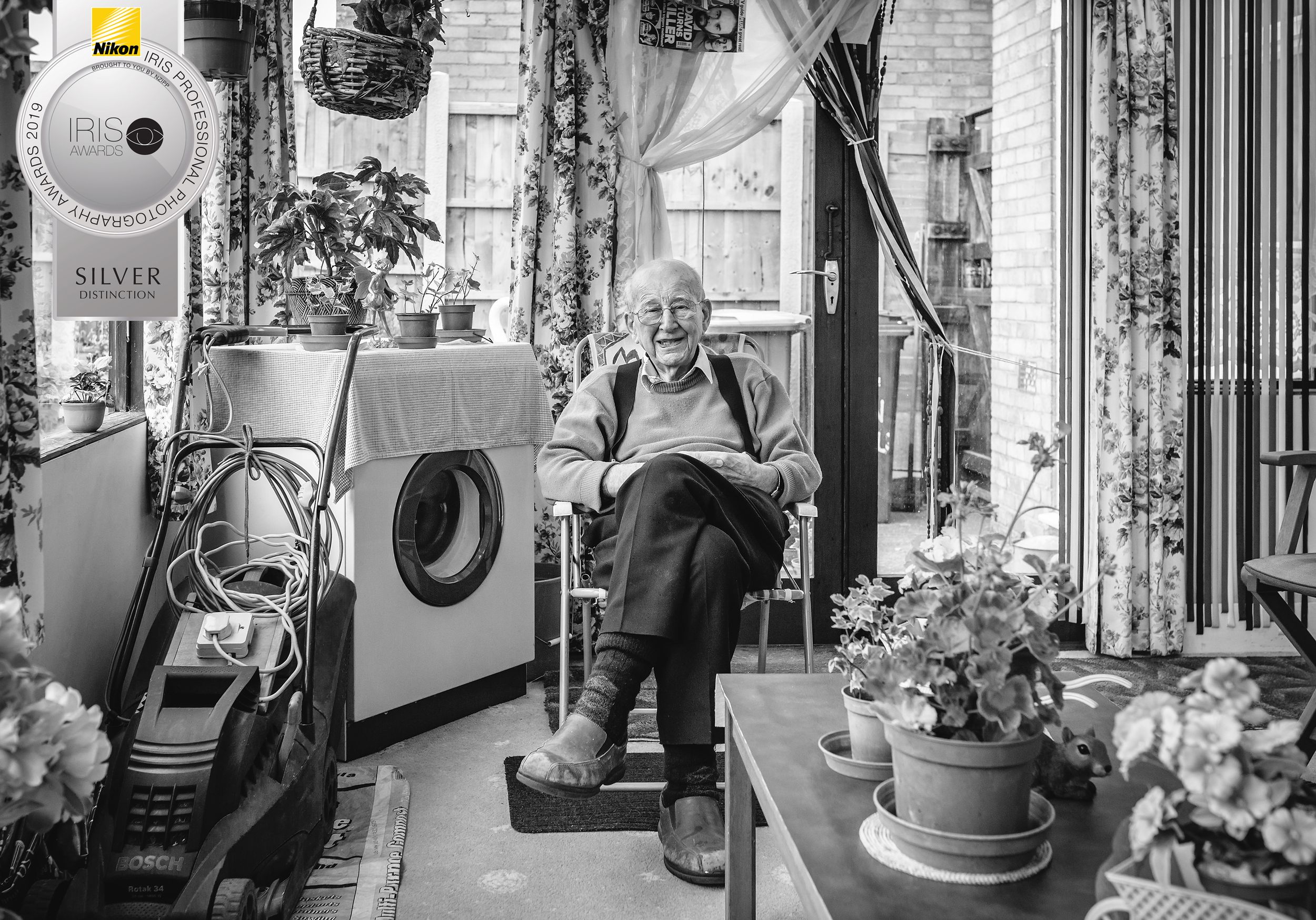 Grandad sitting in a chair in conservatory surrounded by plants, washing machine and lawn mower